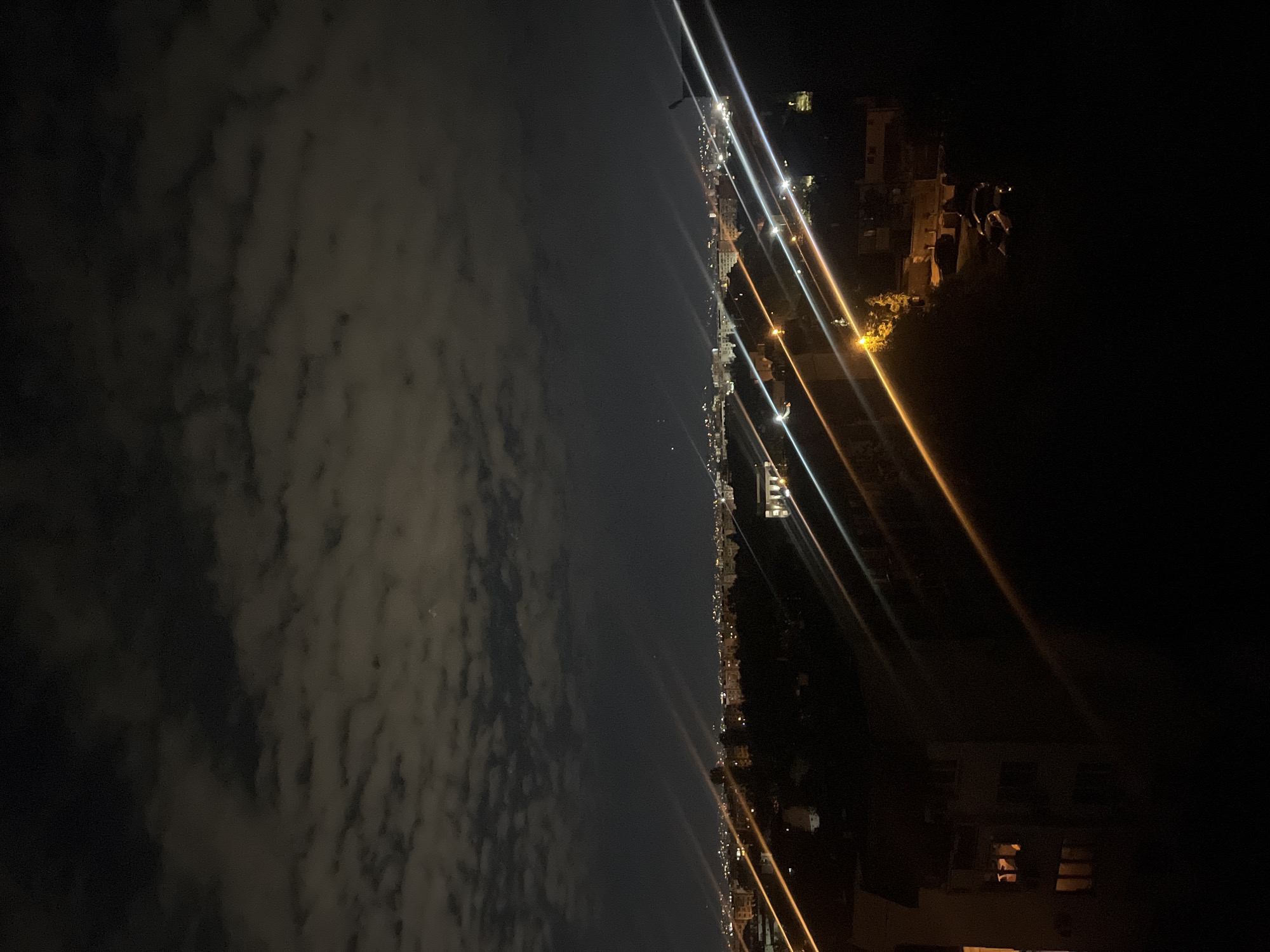 An eye-level, outdoor, nighttime image of a dark scene divided horizontally: the left side is a dark sky with many clouds; the right side is a cityscape with rows of lit buildings and streets. Light beams are visible throughout. The location is likely Sofia, Bulgaria.
