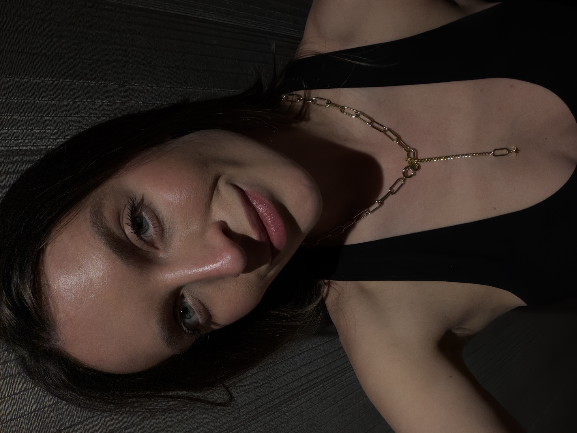A rotated close-up portrait features a person, likely female, with dark hair and light-colored eyes, lying down and looking directly at the camera with a soft, gentle smile. Her face is well-lit on one side, highlighting her skin, subtle makeup, and hydrated lips.
She is wearing a black top and a prominent gold-colored necklace. The necklace is a large paperclip chain design, with a finer extension chain that features a small, delicate star-shaped charm hanging at the center of her chest.
The background is a dark, textured surface, possibly upholstered furniture or bedding, with faint horizontal lines creating a subtle pattern. The lighting is dim and focused, creating a dramatic contrast between the illuminated parts of her face and the shadowed areas of the scene. This directional lighting emphasizes the contours of her face and the metallic gleam of the necklace. The overall mood is intimate and relaxed, suggesting an indoor setting, likely during the evening or night. There is no visible text in the image.
