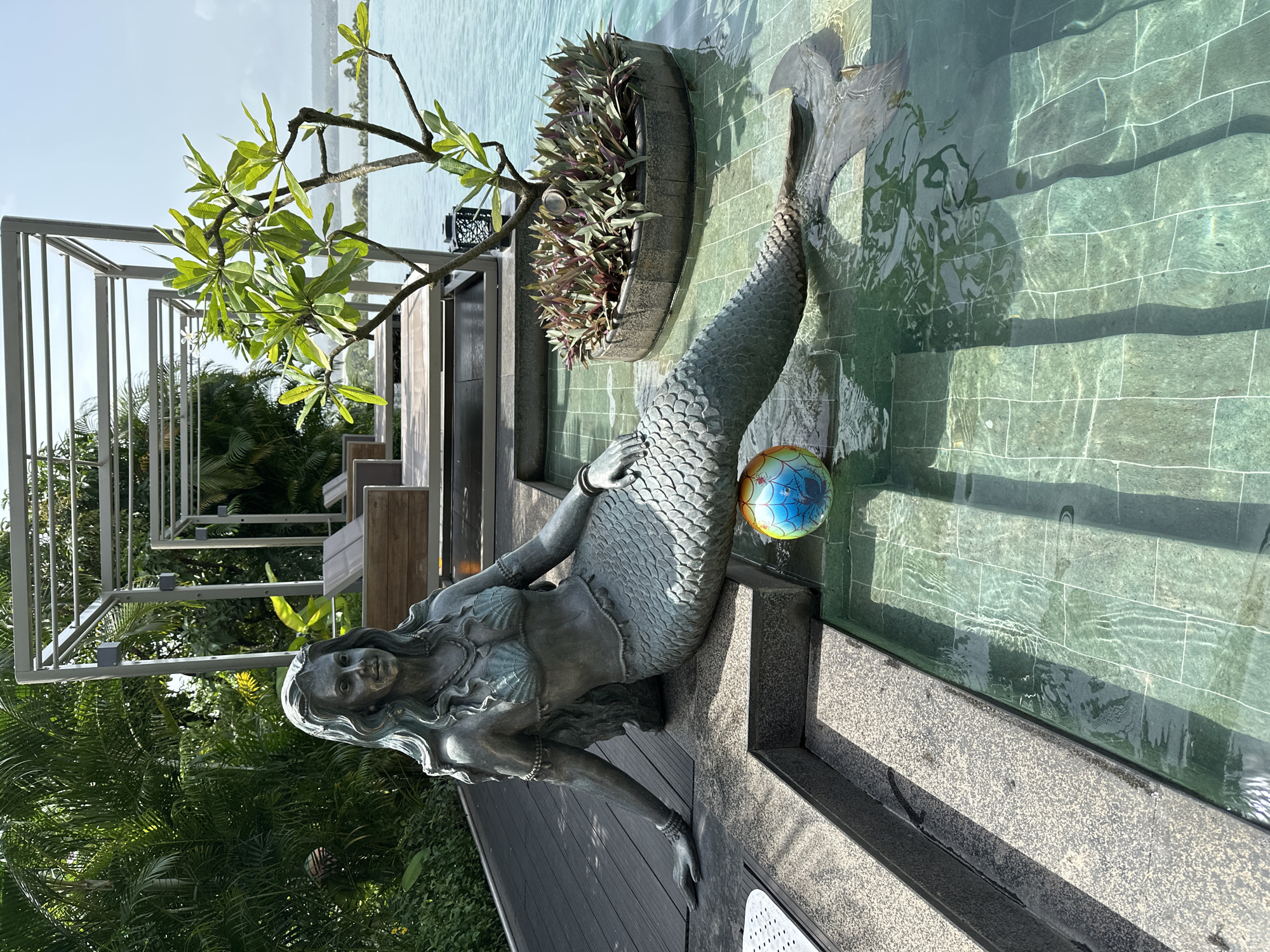 This image depicts a bronze statue of a mermaid partially submerged in a shallow, tiled swimming pool at the Grand Hyatt Goa in Vagator, India. The mermaid is seated on the edge of the pool, with her tail extending into the water. In front of her, a colorful ball with a globe design floats in the pool. The pool area is surrounded by lush greenery, including palm trees and a small decorative plant in a stone planter. In the background, there is a modern outdoor seating area with lounge chairs and a pergola-like structure, overlooking a body of water that appears to be the Arabian Sea. The sky is clear and blue, suggesting a bright, sunny day.