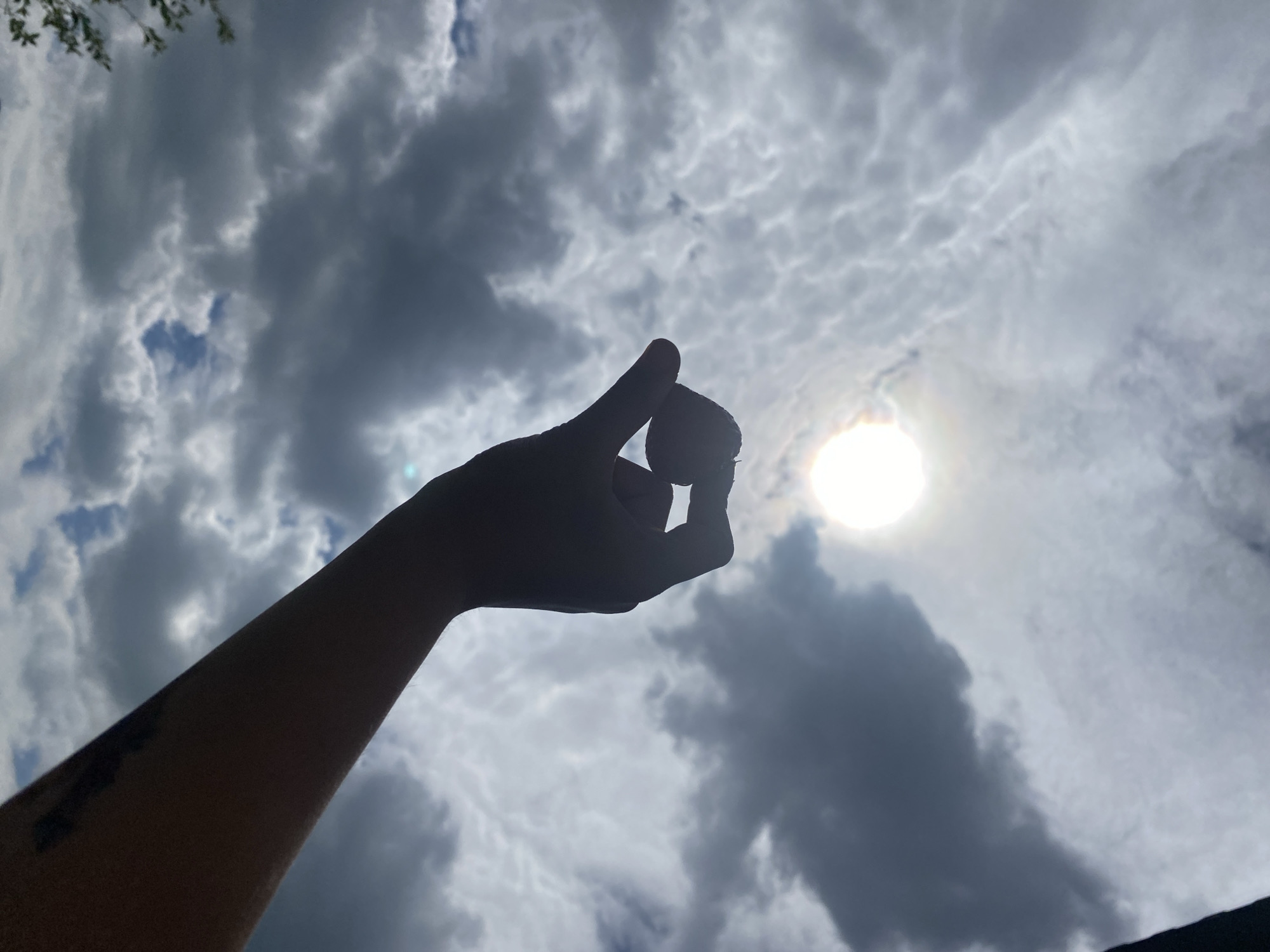 A person's silhouetted forearm and hand are visible against a cloudy sky with the sun shining through. The hand is holding a small, round object, possibly a berry or a small fruit, between their thumb and forefinger. The sky is filled with a mix of dark and light clouds, with patches of blue sky visible in the distance. There is a faint rainbow-like effect around the sun. The image appears to have been taken outdoors, possibly in a park or natural area. The specific location in the city of Tujmazy, Russia, is not discernible from the image itself.