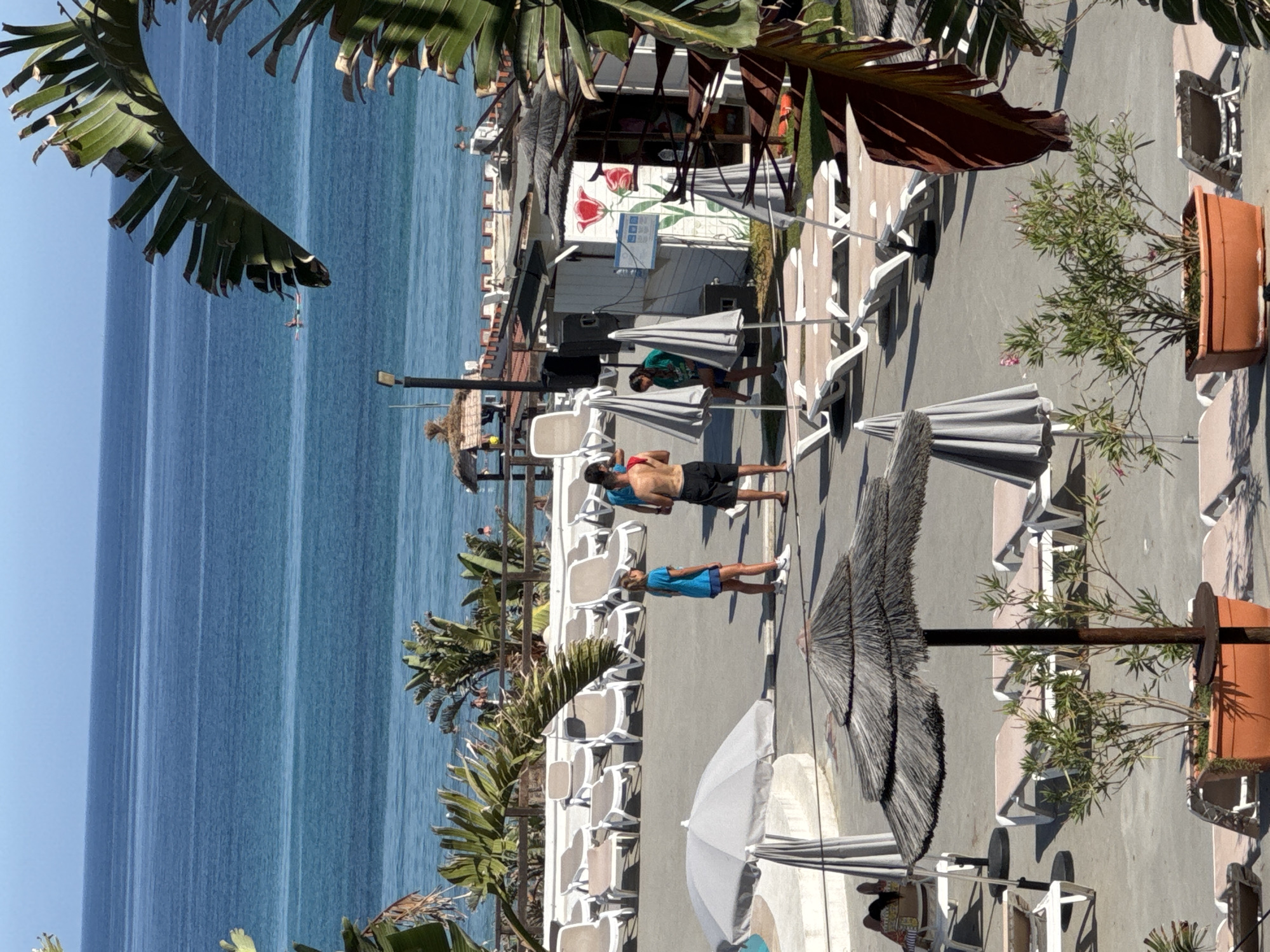 The image depicts a sunny day scene at a coastal resort in Isola delle Femmine, Italy. The foreground is dominated by a paved area with numerous empty lounge chairs arranged in rows. Several thatched umbrellas and modern gray umbrellas provide shade. Potted plants with green foliage are strategically placed throughout the area. In the mid-ground, three individuals are present: a man with his back to the camera wearing black shorts, a young girl in a blue t-shirt and shorts, and a man in a green t-shirt. They appear to be interacting near a small white building adorned with a large red tulip painting. The background features the vast blue Mediterranean Sea, with a single person visible on a paddleboard in the distance. The clear sky suggests good weather conditions. The resort appears to be situated along a coastline with a sandy beach or a promenade visible beyond the lounge chairs.