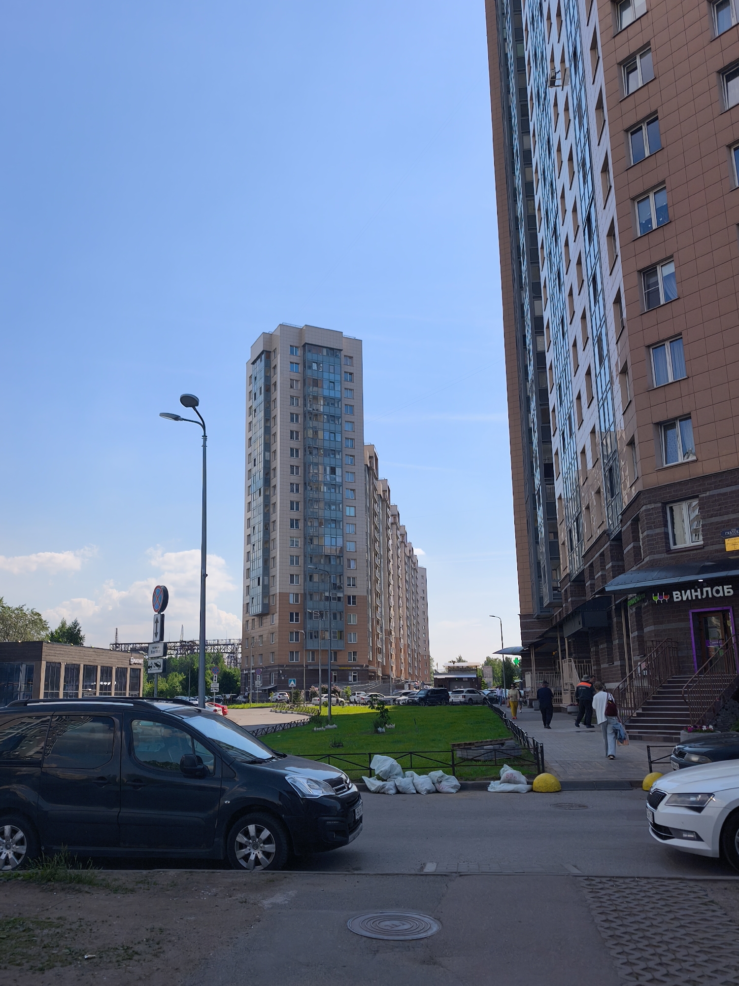 This is a street-level, eye-level, medium shot of a residential area in Saint Petersburg, Russia. The scene captures a row of modern apartment buildings under a clear blue sky. The apartment buildings are tall, multi-story structures with contemporary architectural designs, featuring a mix of light-colored facades and large glass windows. On the right side of the frame, a larger building with a brown tiled facade dominates the foreground, with the entrance to a shop labeled "ВИНЛАБ" (Vinlab) visible. On the left, a narrower, taller apartment building is visible, with a streetlight and traffic signs in front of it.
In the foreground, a black minivan is parked along the curb. The street appears to be paved with asphalt. Several white bags, presumably trash or construction debris, are piled on the side of the road, along with two yellow bollards. A few people are walking on the sidewalk in front of the Vinlab store, one individual wearing a white top and blue jeans, another in a dark suit, and a person in an orange safety vest. A white car is partially visible on the right side of the image.
The environment suggests a developed urban setting with residential buildings, commercial establishments, and infrastructure for transportation and public services. The presence of green grass areas between buildings indicates some landscaping. The time of day appears to be daytime, likely late morning or early afternoon, given the bright sunlight and the clear sky.