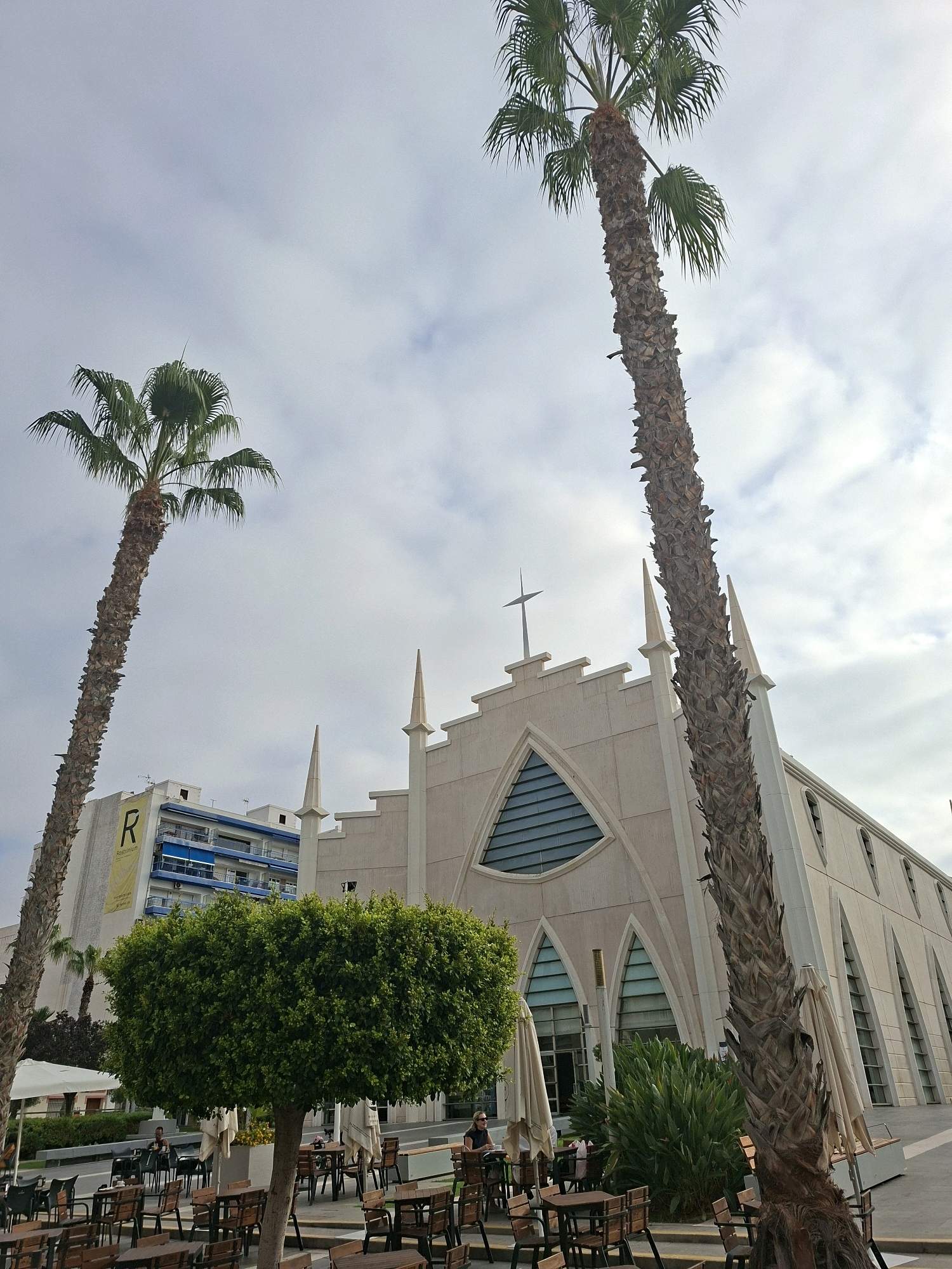 The image depicts an outdoor seating area with multiple tables and chairs, set against the backdrop of a modern church and a tall building. Two large palm trees are prominent in the foreground, one on the left and another to the right, with its trunk obscuring a significant portion of the church's facade. The church, with its distinctive pointed arch windows and a cross atop its roof, appears to be constructed from a light-colored material, possibly stone or stucco. To the left of the church, a multi-story building with blue balconies and a large yellow sign bearing the letter "R" is visible. The seating area is partially shaded by umbrellas and features lush green foliage, including a large, rounded tree in the middle ground. Several people are seated at the tables, engaged in conversations or dining, suggesting a lively atmosphere. The sky is overcast, with scattered clouds. This scene is located in Torrevieja, Spain, specifically in front of the Virgen del Carmen Parish Church, which is known for its modern architectural style.