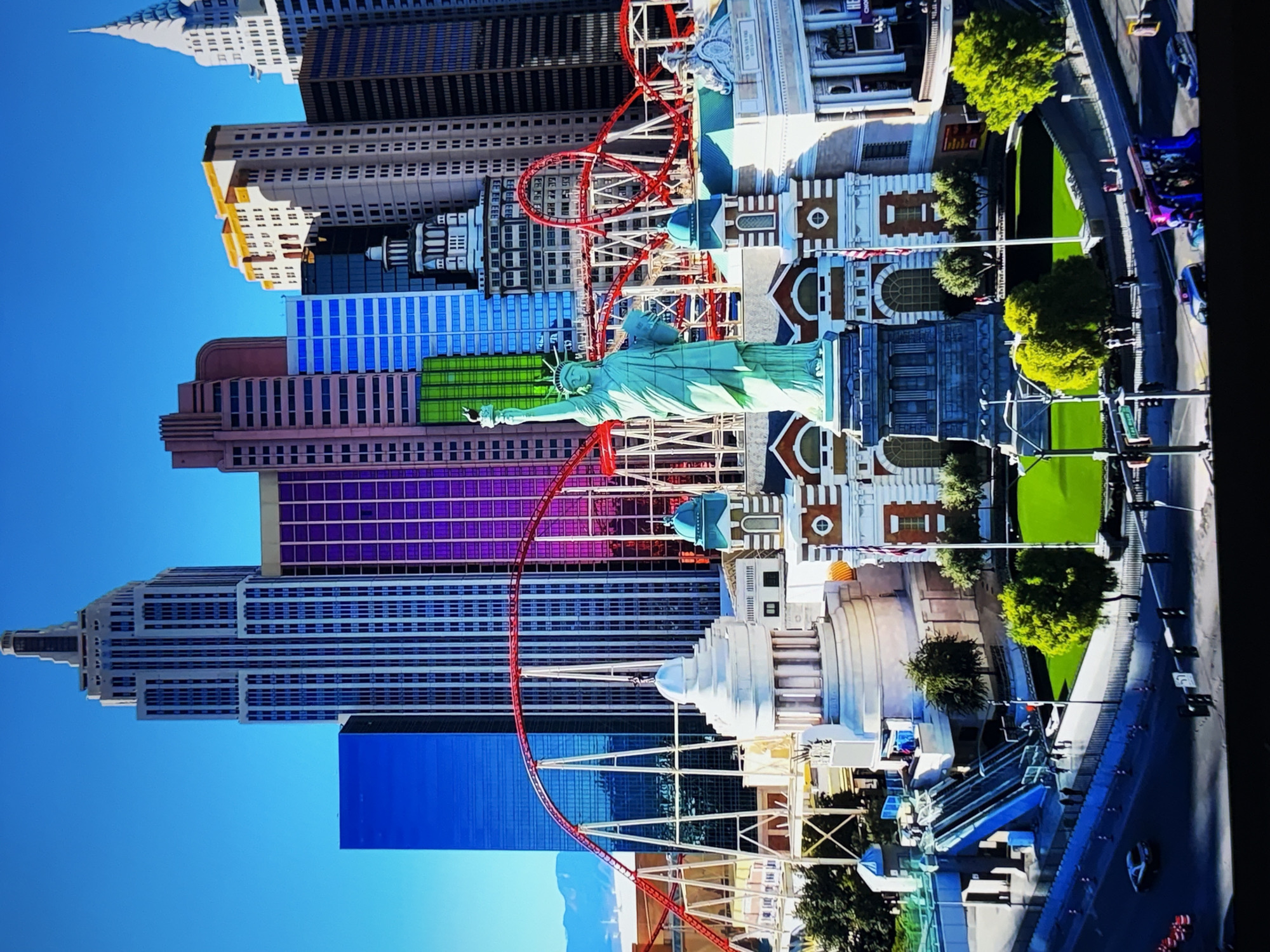 The image depicts a vibrant cityscape with a central focus on a replica of the Statue of Liberty, surrounded by various New York City-inspired architectural landmarks, including skyscrapers and a roller coaster. The buildings are a mix of modern designs with colorful facades, such as blue and purple glass, and more traditional stone structures. A red roller coaster snakes its way through the scene, adding a dynamic element. The Statue of Liberty itself stands prominently on a pedestal. In the foreground, there is a grassy area, a street with traffic, and pedestrian walkways with escalators. The sky is a clear, bright blue, suggesting a sunny day. There are some people visible on the walkways and a few cars on the street. The overall impression is of a themed entertainment complex, likely a casino or resort, designed to evoke the atmosphere of New York City. The image does not depict the city of Burgas, Bulgaria, but rather a representation of New York City landmarks.