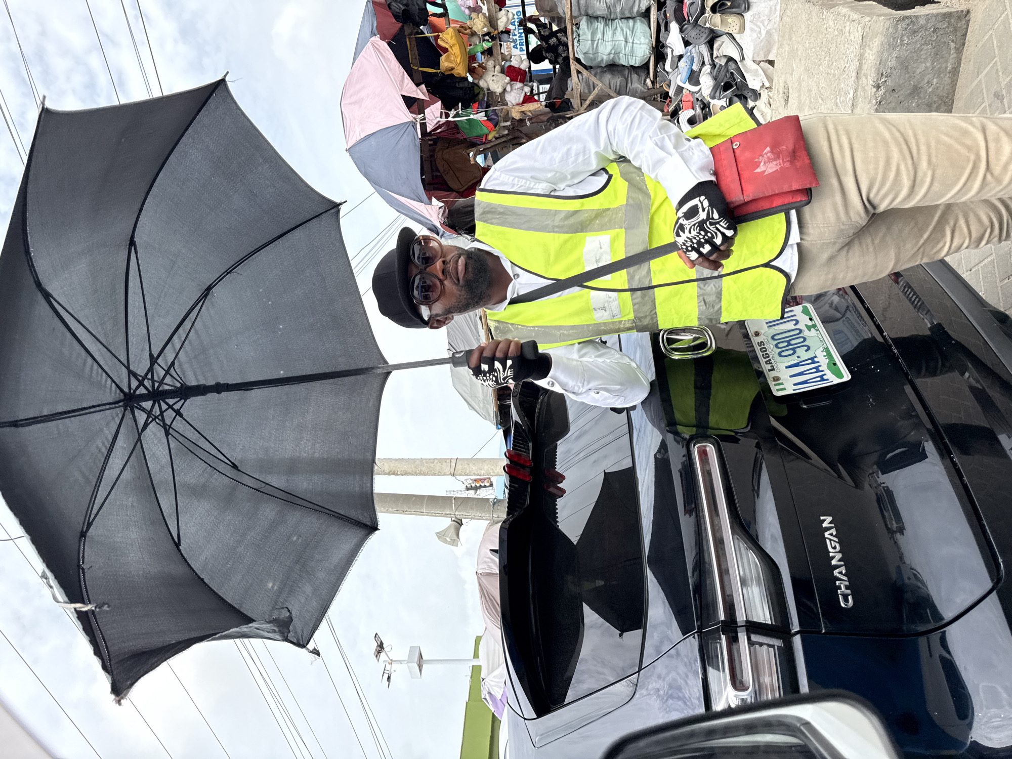 A man in a black hat, sunglasses, white shirt, yellow reflective vest, beige pants, and skull gloves holds a black umbrella. He stands next to a black Changan car with a green and white license plate that reads "LAGOS LAA 980 JC". The background includes a market scene with various items, including shoes, umbrellas, and stacked materials. The setting appears to be a street in Ilado, Nigeria, based on the license plate.