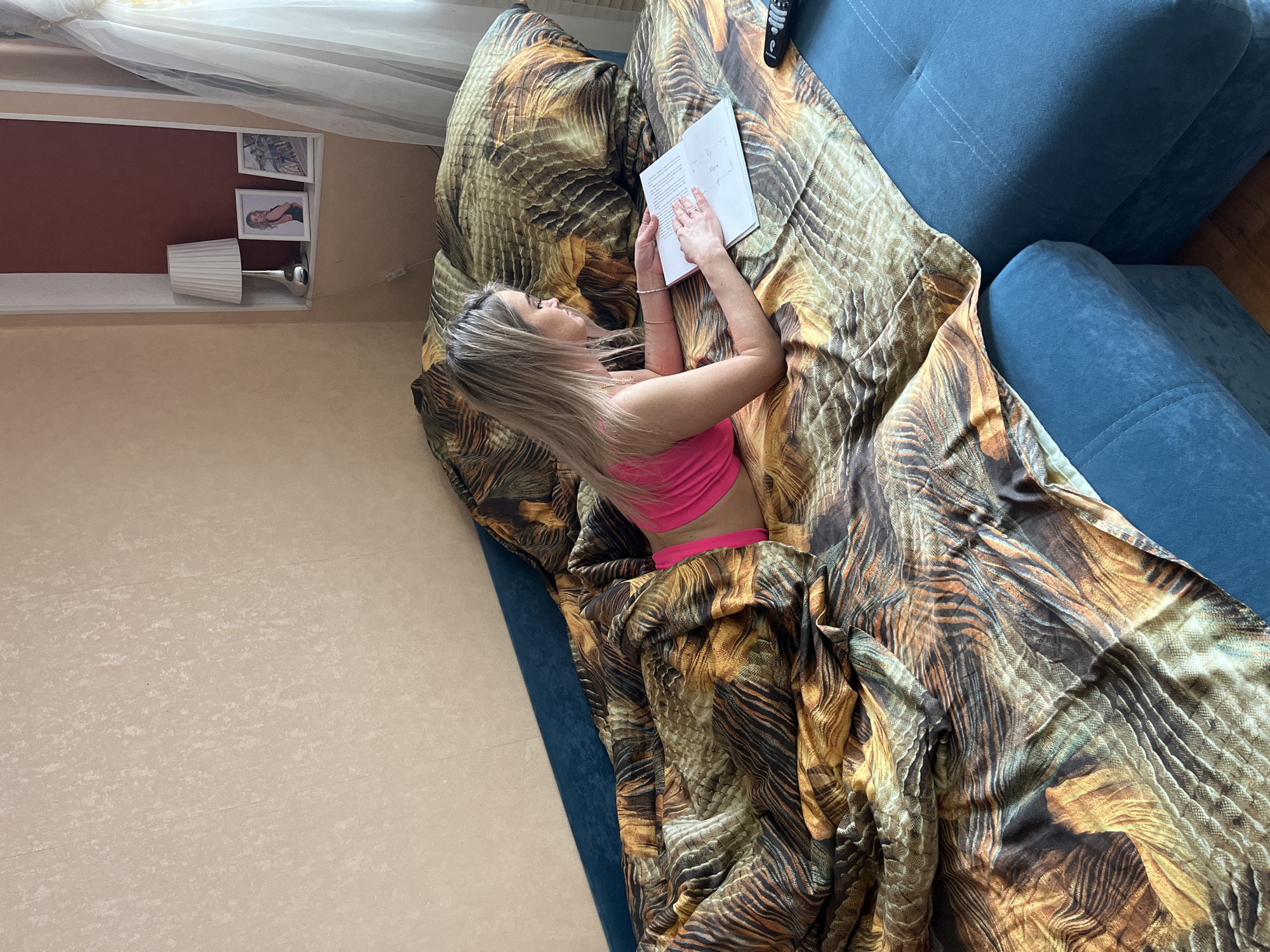 A single female individual is depicted lying prone on a blue upholstered sofa-bed in an interior room located in Chaikovsky, Russia. The female has light blonde hair and is wearing a bright pink top and light-colored bottoms. She is positioned on her stomach, resting on her elbows, and is holding an open book or document with both hands, appearing to read its contents. Her left index finger rests on the page.
The sofa-bed is covered by a duvet featuring a distinct brown, orange, yellow, and black feathered or scaled pattern. To the right of the female, on the blue sofa cushion, a black television remote control is visible. The wall behind the sofa is covered in light beige textured wallpaper. To the upper right, a recessed architectural niche is present. Inside this niche, against a dark reddish-brown back wall, are a white pleated table lamp with a silver base, and two framed photographs. One photograph displays a woman in a black outfit, while the other shows an outdoor scene. A sheer white curtain hangs on the far right, partially obscuring a window.