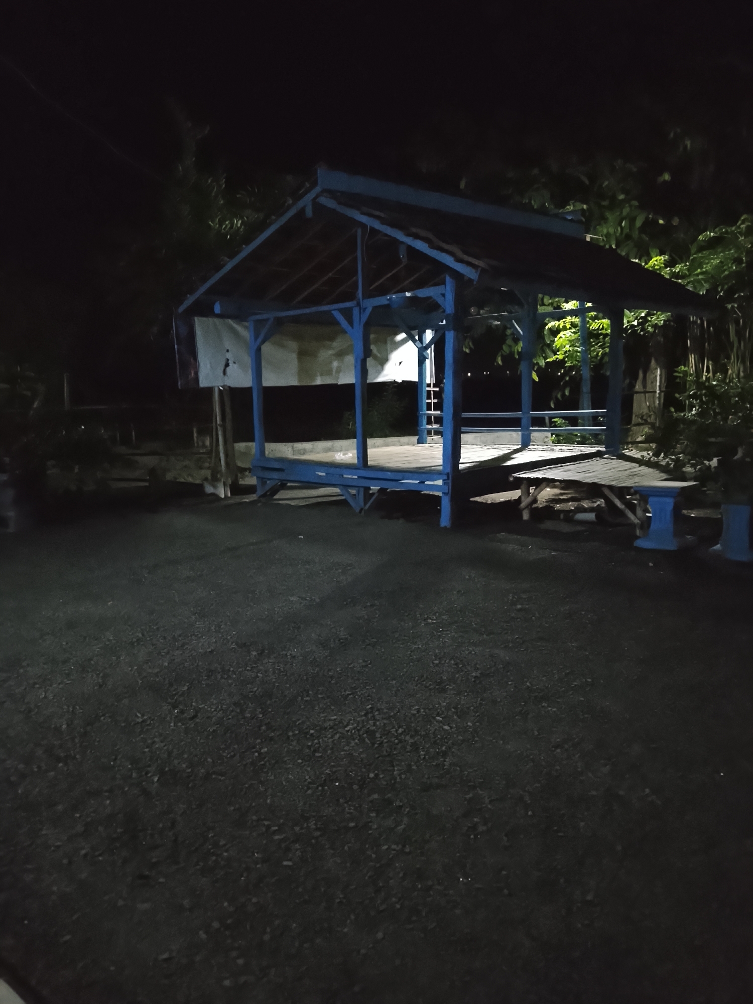 The image depicts a blue, open-sided wooden pavilion at night, possibly located in Dawuan, Indonesia. The pavilion has a pitched roof and a wooden floor, with a small wooden table and bench set up within its confines. The surrounding area is dark, with some vegetation visible behind the pavilion. The ground in front of the structure appears to be paved with dark gravel. There are no people visible in the image.