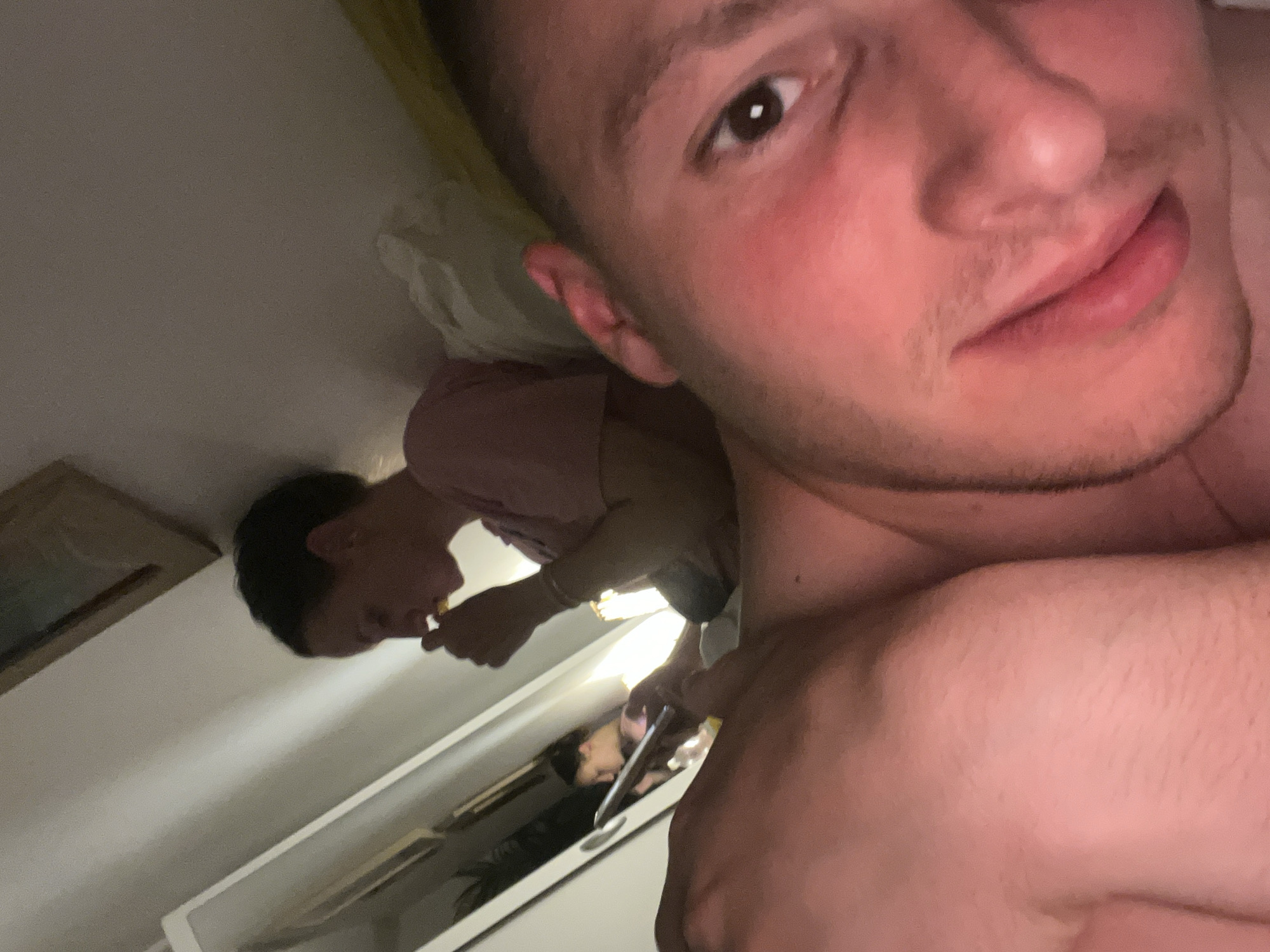The image, rotated 90 degrees counter-clockwise from its likely original orientation, features a close-up selfie taken indoors, contextualized to be in Vitry-sur-Seine, France. The primary subject is a young man whose face occupies the majority of the foreground, specifically the right side of his face from chin to forehead. He looks directly at the camera with dark brown eyes and a neutral to slightly smiling expression. His fair skin shows some flushing on the cheeks and light stubble on his chin and upper lip. Part of his bare shoulder or upper arm is visible in the lower right, suggesting he is shirtless or wearing minimal upper body attire.
In the background, a reflection in a mirror or polished surface reveals another person with dark hair, wearing a light-colored (possibly pink) short-sleeved top. This individual appears to be holding a yellow or orange item to their mouth, as if eating. A bright artificial light source is visible in the reflection, positioned between the two figures. The reflection also shows a white surface, likely a table or dresser, and a framed picture on the wall to the left of the reflected person. The overall dimness with artificial lighting suggests an evening or nighttime setting within a domestic space, likely a bedroom. No discernible text is visible in the image.