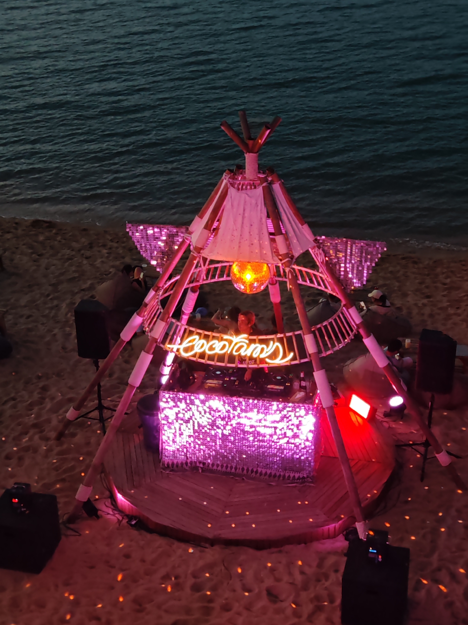 This is an aerial, high-angle shot of an outdoor DJ setup on a sandy beach at night. The DJ booth, covered in shimmering pink sequins, is the central focus. Above the booth, a teepee-like structure is constructed from wooden poles and draped with fabric, adorned with a disco ball. Neon lights spelling "Cocotamy's" in script font illuminate the DJ booth. To the sides of the DJ booth are large black speakers. In the background, the ocean is visible, with gentle waves lapping the shore. Scattered across the sand are small, glowing orange lights, likely from other decorative elements or ambient lighting. Several beanbag chairs are placed around the setup, some occupied by individuals who appear to be attendees, while others are empty. One person is visible behind the DJ booth, appearing to be the DJ. The overall ambiance suggests a lively beach party or event. The location is identified as Baan Bo Phut, Thailand.