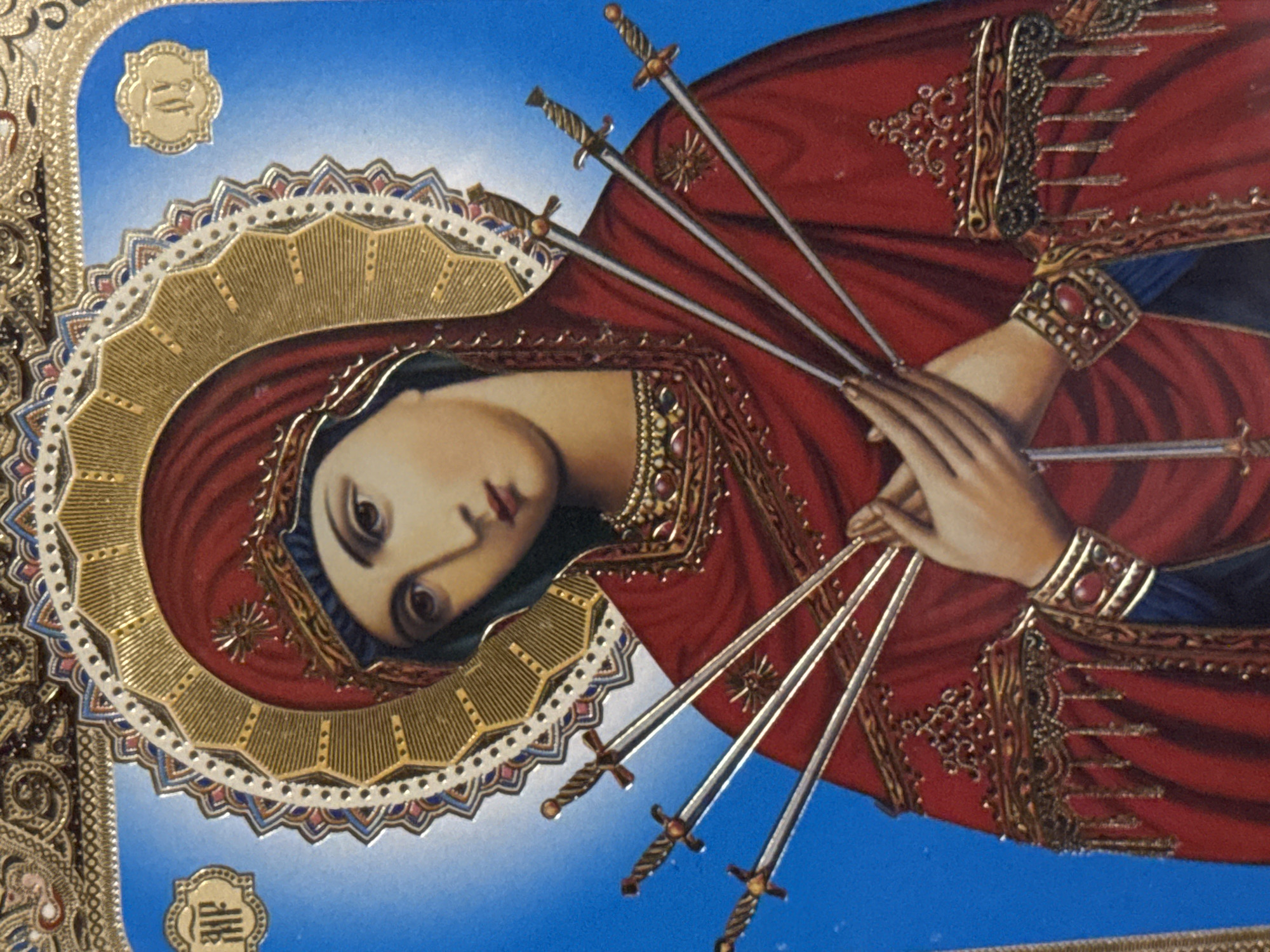 This is a close-up, full-frame, portrait-style image of an icon depicting the Virgin Mary. The Virgin Mary is shown from the shoulders up, wearing a red veil and garment. She has a halo around her head, which is intricately decorated with gold and white patterns. Seven swords are depicted piercing her chest, with their hilts visible and blades extending outwards. Her hands are clasped together over the swords. The background is a bright blue, with ornate gold embellishments at the top and two small, circular gold medallions with Greek letters on either side of her head. The image is detailed and appears to be a religious artifact, likely an icon of Our Lady of Sorrows. The location is unknown.