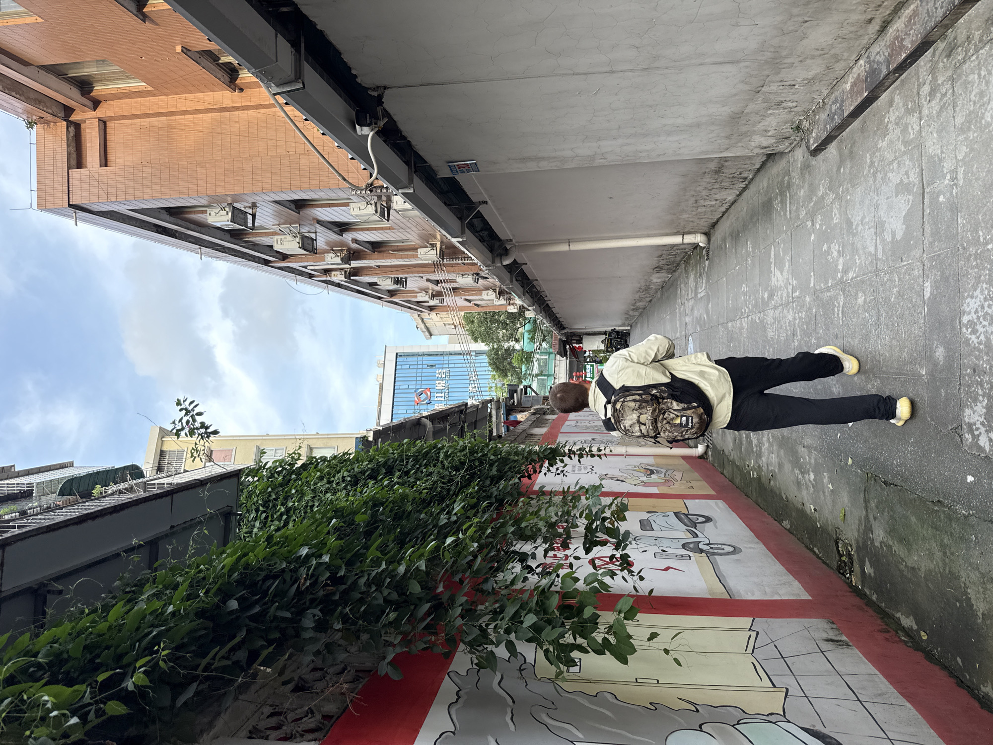 A pedestrian walks through a narrow alley in the Haizhu District, China. The alley is bordered on one side by a tall building with multiple air conditioning units and on the other side by a wall covered in vines and a cartoon mural. The alley is paved with grey tiles and the sky is partly cloudy. The pedestrian is wearing black pants, a light-colored jacket, yellow shoes and a backpack.