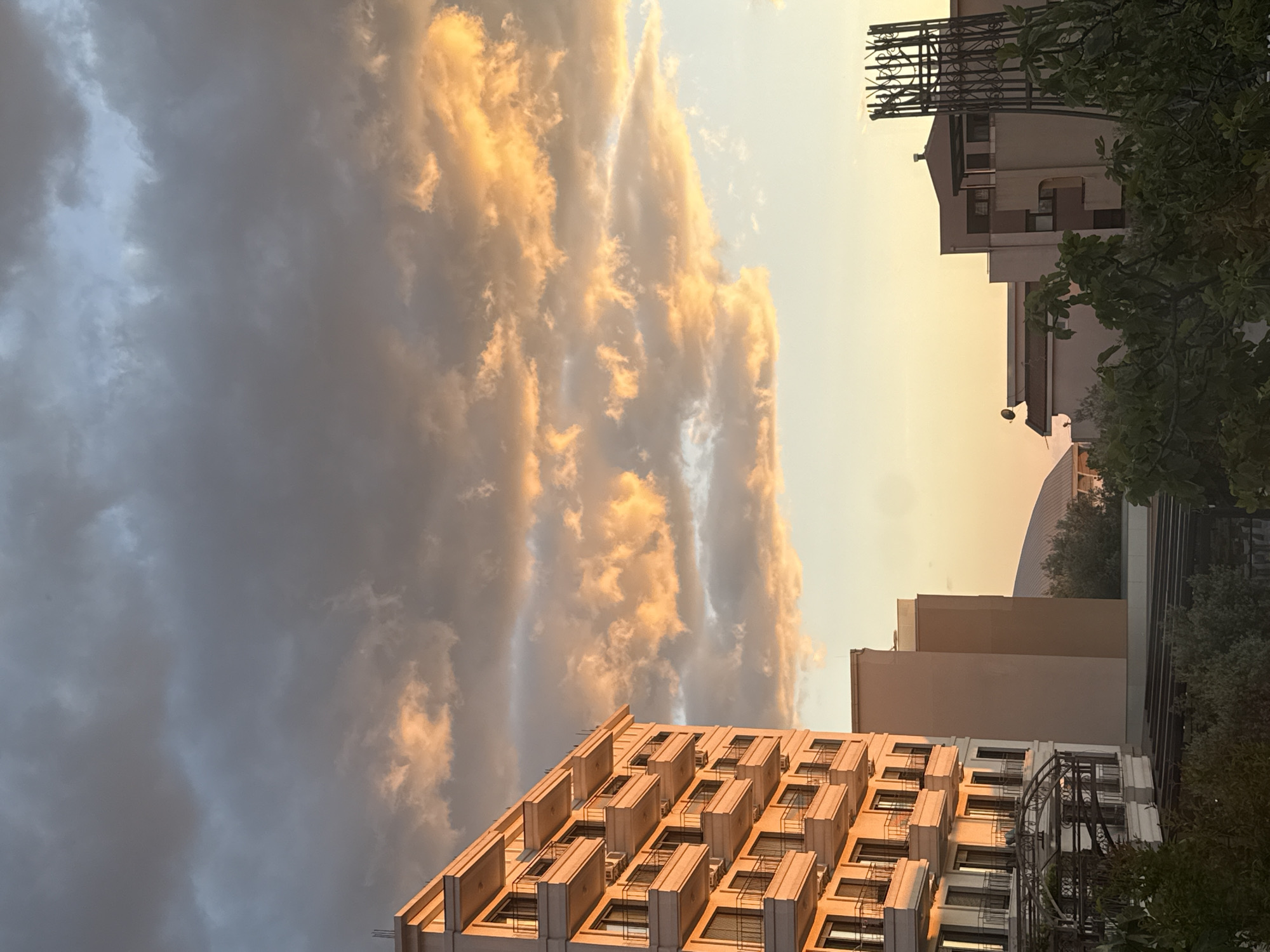 The image captures a dramatic urban landscape at either sunrise or, more likely, sunset, with striking atmospheric conditions over Bar, Montenegro. The sky is a prominent feature, with heavy, dark grey storm clouds dominating the left side, contrasting sharply with brightly illuminated, golden-orange clouds on the right, bathed in the low sun's warm light. The sky transitions to a pale, clear yellow-orange on the far right.
In the foreground, a modern, light-colored building with a repetitive pattern of rectangular balconies or window structures reflects the golden light. Behind it, on the right, stands a darker building with an ornate black metal structure on its roof, partially obscured by lush green trees and bushes. Other buildings with beige facades are visible in the mid-ground, leading to distant mountains or hills. A large, curved architectural structure also appears in the upper mid-ground.
The time of day is clearly indicated by the intense golden and orange hues. The juxtaposition of dark and brightly lit clouds suggests dynamic weather, possibly after a storm has passed or during a transient period of dramatic cloud formation. There is no discernible human activity or visible text in the scene.