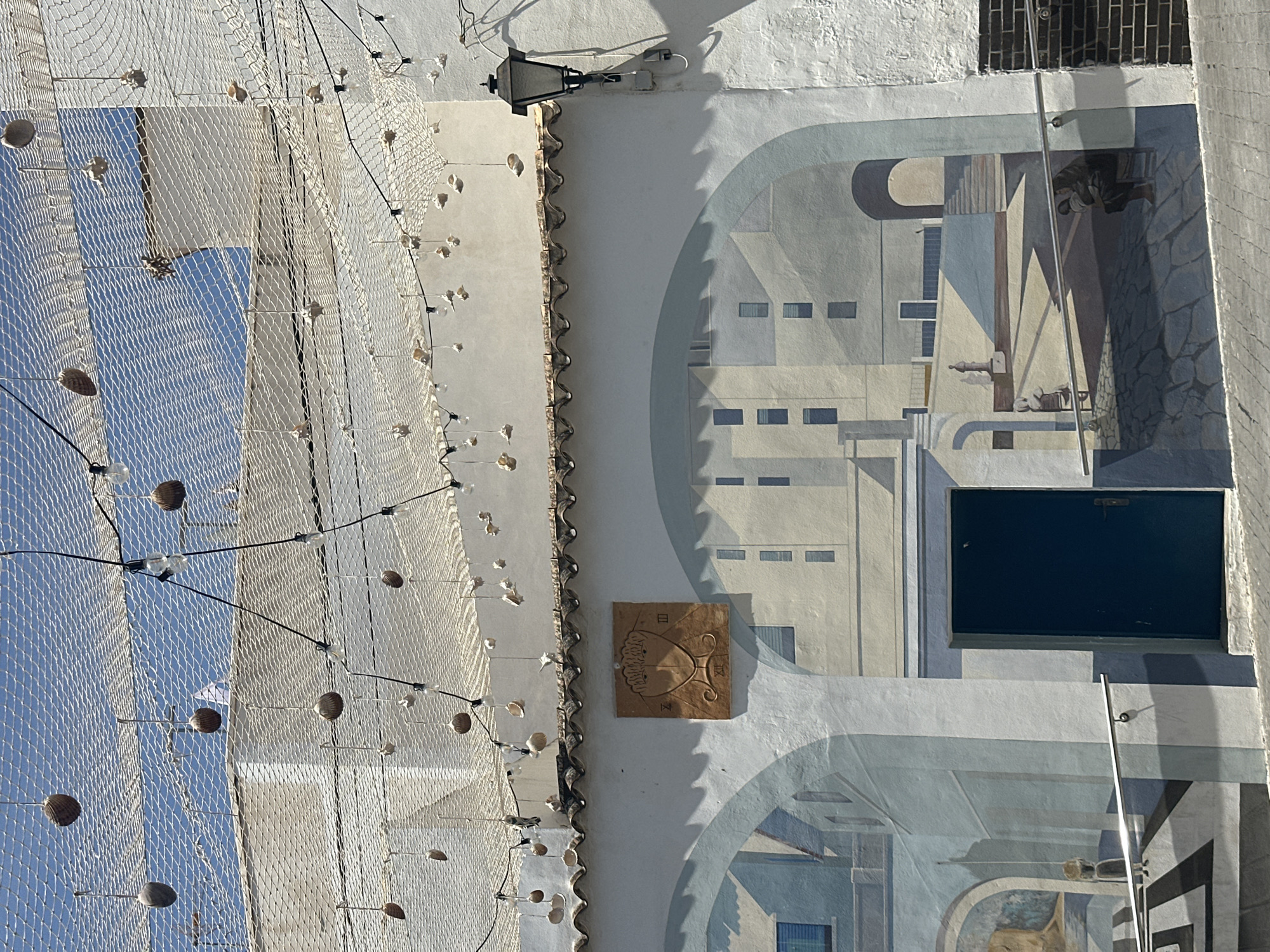 The image captures an architectural facade in Calp, Spain. The scene features a white building with an artistic mural depicting various architectural elements in grayscale tones. A black rectangular door is visible within the mural. To the left, a decorative fishing net hangs, adorned with small, round objects resembling seashells or lights, suspended by thin black wires. Above, a wrought-iron street lamp is affixed to the building. The sky, a light blue, is visible through the fishing net. A square sundial is mounted on the wall.