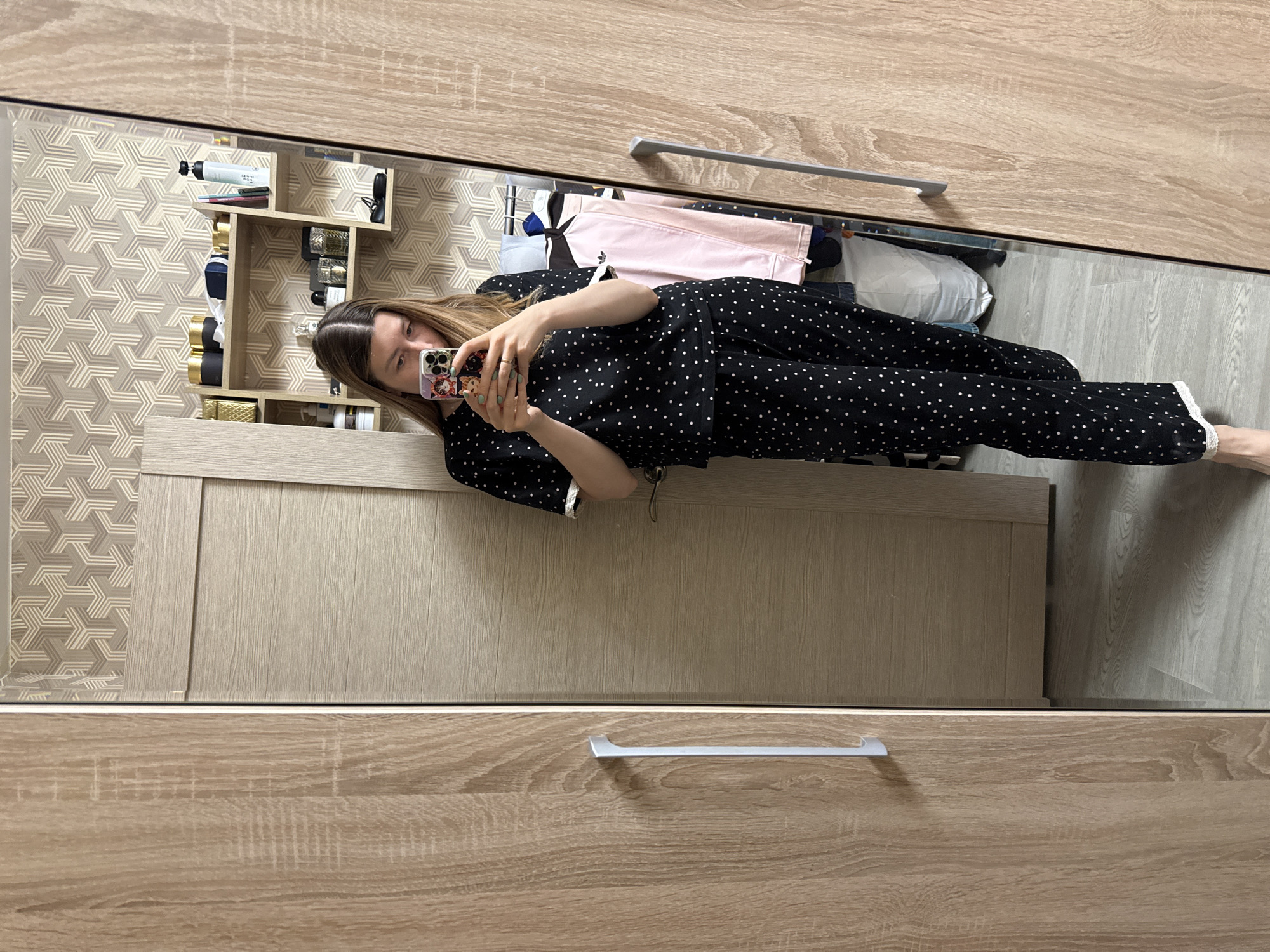 A woman is visible in a mirror, taking a selfie with her phone. She is wearing black pajamas with white polka dots. The reflection shows a portion of a room with light wood-grain cabinets or drawers, a geometric patterned wallpapered section, and a shelf with various objects including bottles. The location cannot be determined as being in Moscow, Russia, from this image alone.