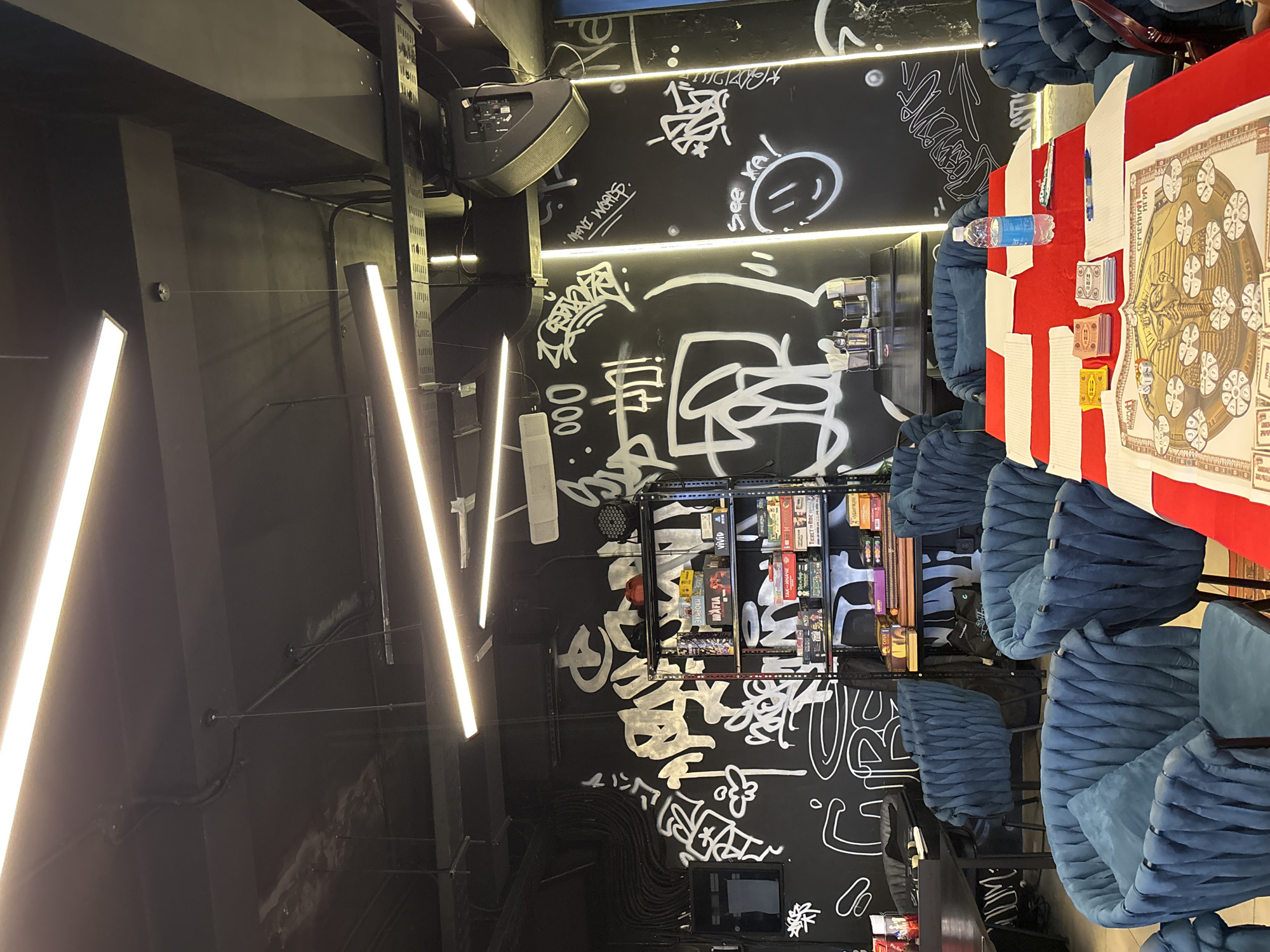 This image captures an indoor setting, likely a board game cafe or lounge, characterized by a dark, modern aesthetic. The scene is dominated by black walls heavily adorned with white graffiti and stylized script, giving it an urban, edgy feel. Bright, linear LED strip lights illuminate the space from above, casting a warm glow.
On the right side of the frame, several plush, blue velvet-like chairs with a ribbed design are arranged around a long table. This table is covered with a vibrant red tablecloth, upon which a large, ornate game board with circular motifs and text in Cyrillic ("СЕМЕЙНАЯ ИГРА" - Family Game) is laid out. Adjacent to the game board are playing cards, a plastic water bottle, a pen, and a small notebook, suggesting an ongoing or即将开始 game session.
Against the back wall, a metal shelving unit displays a collection of board games with visible titles such as "TICKET TO RIDE", "VIVID", and "MAFIA". Above the shelving, a speaker is mounted. Additional details include visible wiring and ventilation ducts along the ceiling and walls.
Visible text in the graffiti includes "MINI WORDS.", "See XA!", "000", and what appears to be "Виховихе" (Vikhovikhe) in Cyrillic. The overall ambiance suggests an evening or nighttime setting due to the reliance on artificial lighting. Based on the provided context, this establishment is located in Kuta Selatan, Indonesia. No individuals are visible in the image.