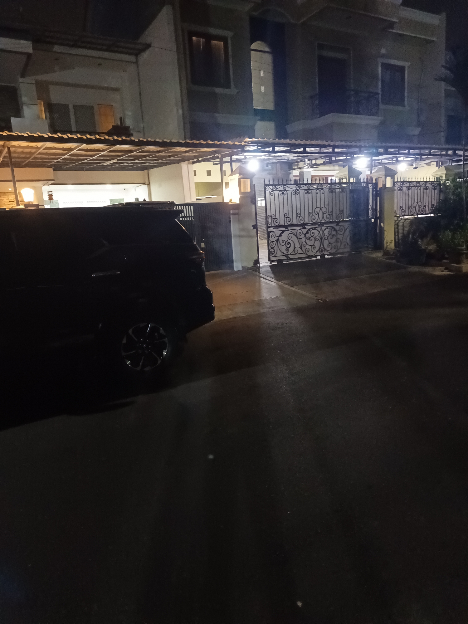 This image captures a nighttime view of a house with a black SUV parked in the driveway, located in the city of North Jakarta, Indonesia. The house appears to be a multi-story building with an attached carport, illuminated by several exterior lights. A black metal gate with an ornate design is visible in front of the property, with one section open. The SUV is a dark color and is partially obscured by shadow. The driveway and surrounding area are paved, and the overall lighting suggests it is nighttime.