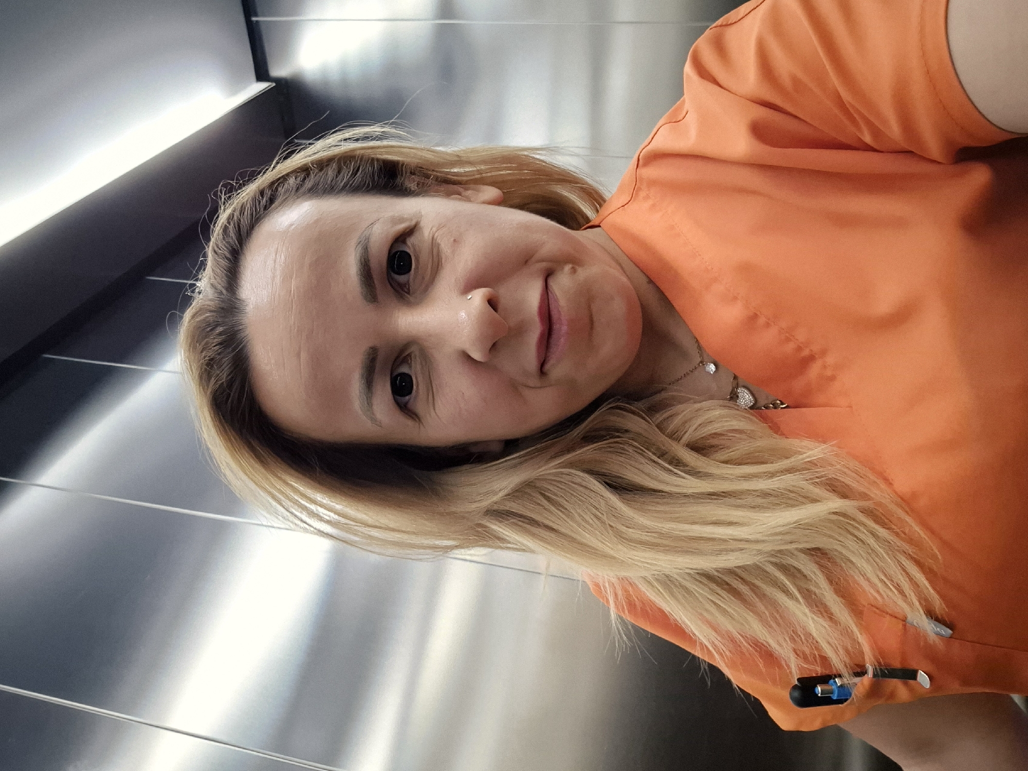 A woman with blonde hair and dark eyes, wearing an orange scrub top, poses for a selfie in what appears to be an elevator. She has a small nose ring and a delicate necklace with a heart-shaped pendant. A blue pen is clipped to her left breast pocket. The elevator walls are metallic and reflective, with vertical lines and overhead lighting visible. The overall lighting suggests an indoor, potentially sterile environment. The location is described as Unknown, Unknown.