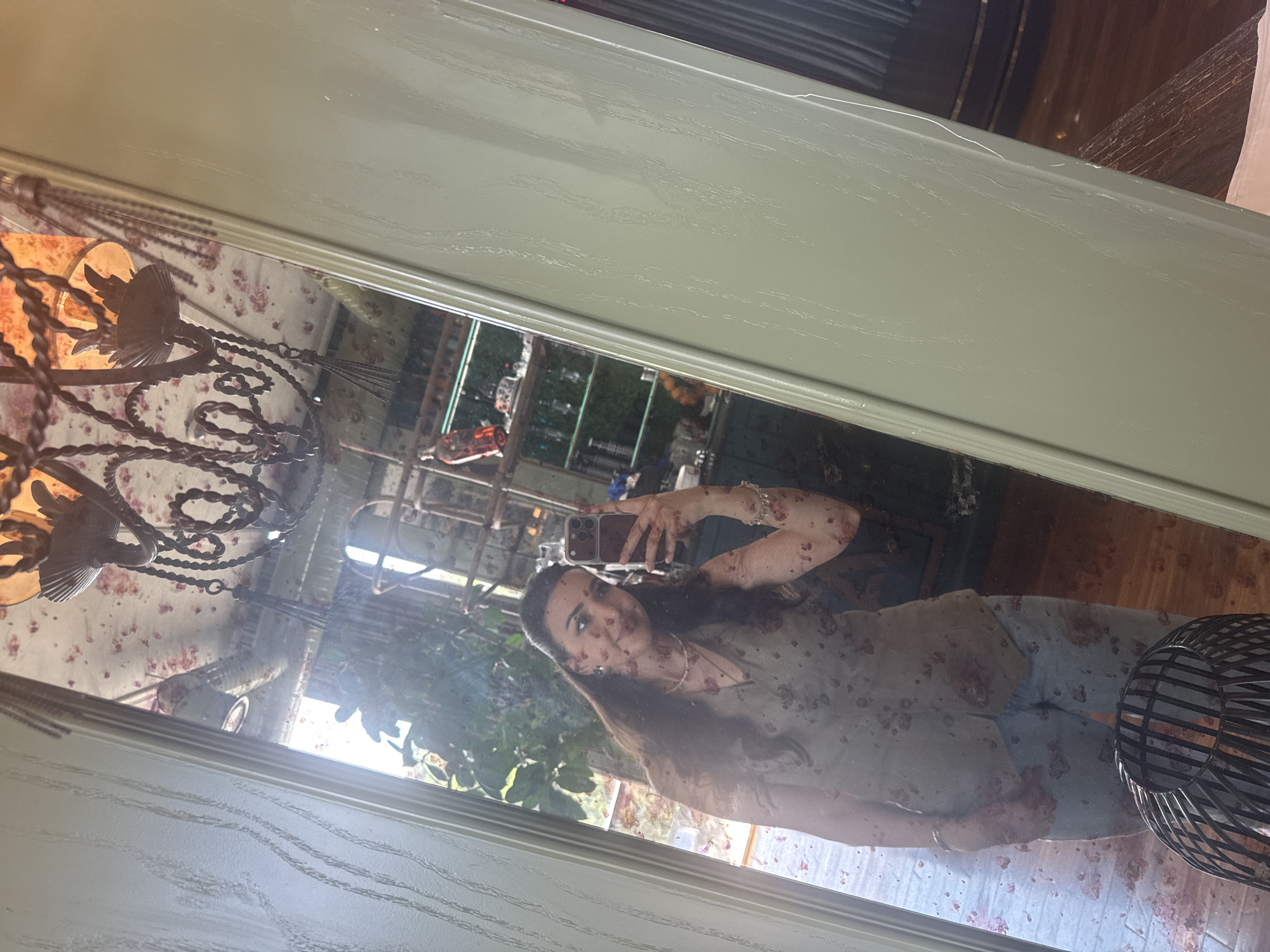A young woman with dark hair tied back, wearing a sleeveless patterned vest over a pearl necklace and light-wash jeans, takes a mirror selfie. She holds a smartphone in her right hand, capturing her reflection. The mirror's surface is smudged, obscuring some details of the room behind her. In the reflection, a chandelier with a floral patterned shade hangs from a patterned ceiling. Behind the woman, shelves are visible, holding bottles and decorative items. The floor appears to be wooden. The image is taken indoors, likely in a residential setting or a boutique.