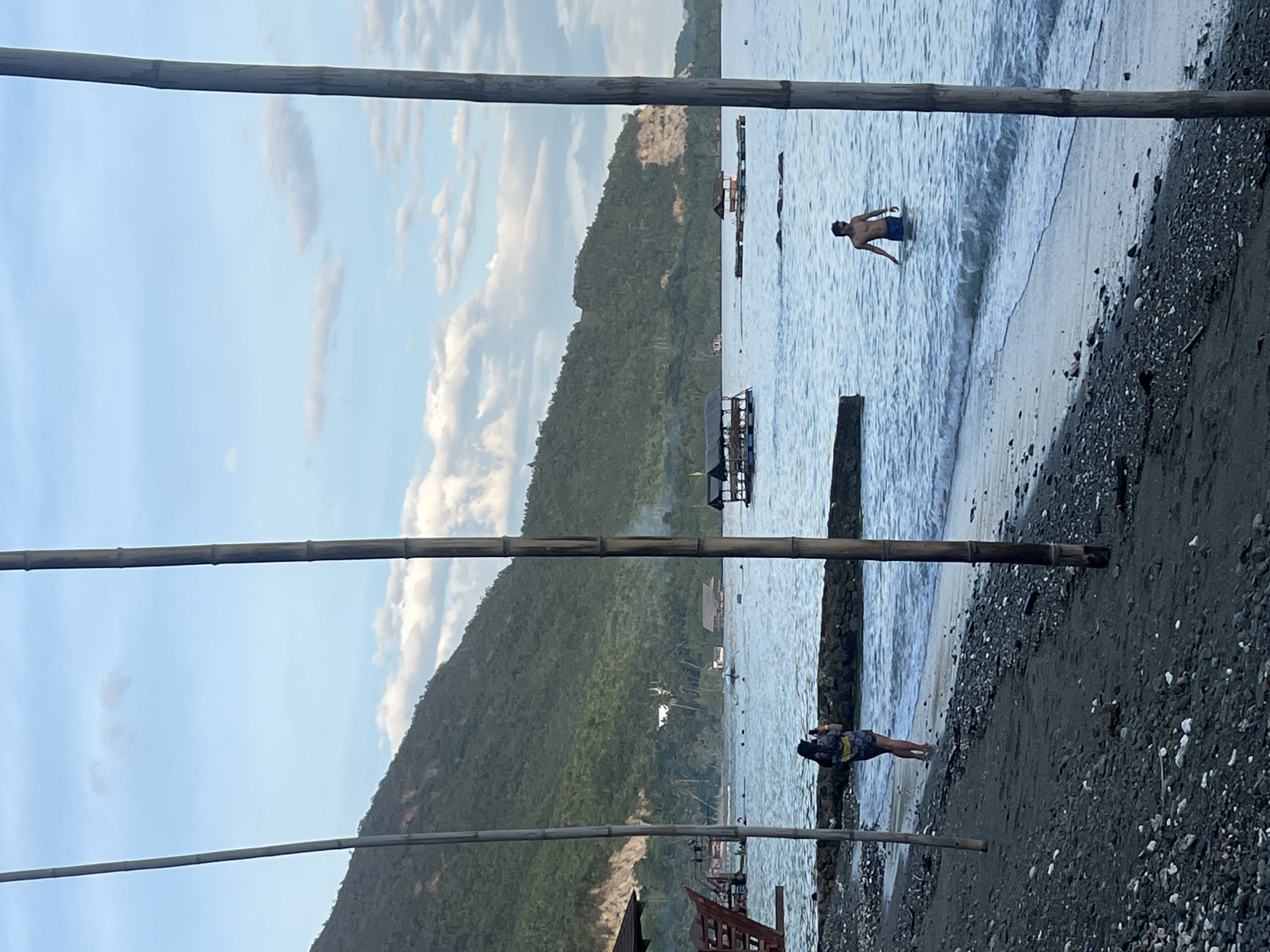 The scene depicts a beach in Banaybanay, Philippines. The image is framed by three horizontal bamboo poles. In the upper right of the frame, a man is wading in the ocean. Further down, a woman is lying on the beach. The beach is dark, with a mixture of sand and small rocks. In the background is a green mountain covered in vegetation. There are also several floating structures in the water. The sky is partially cloudy.