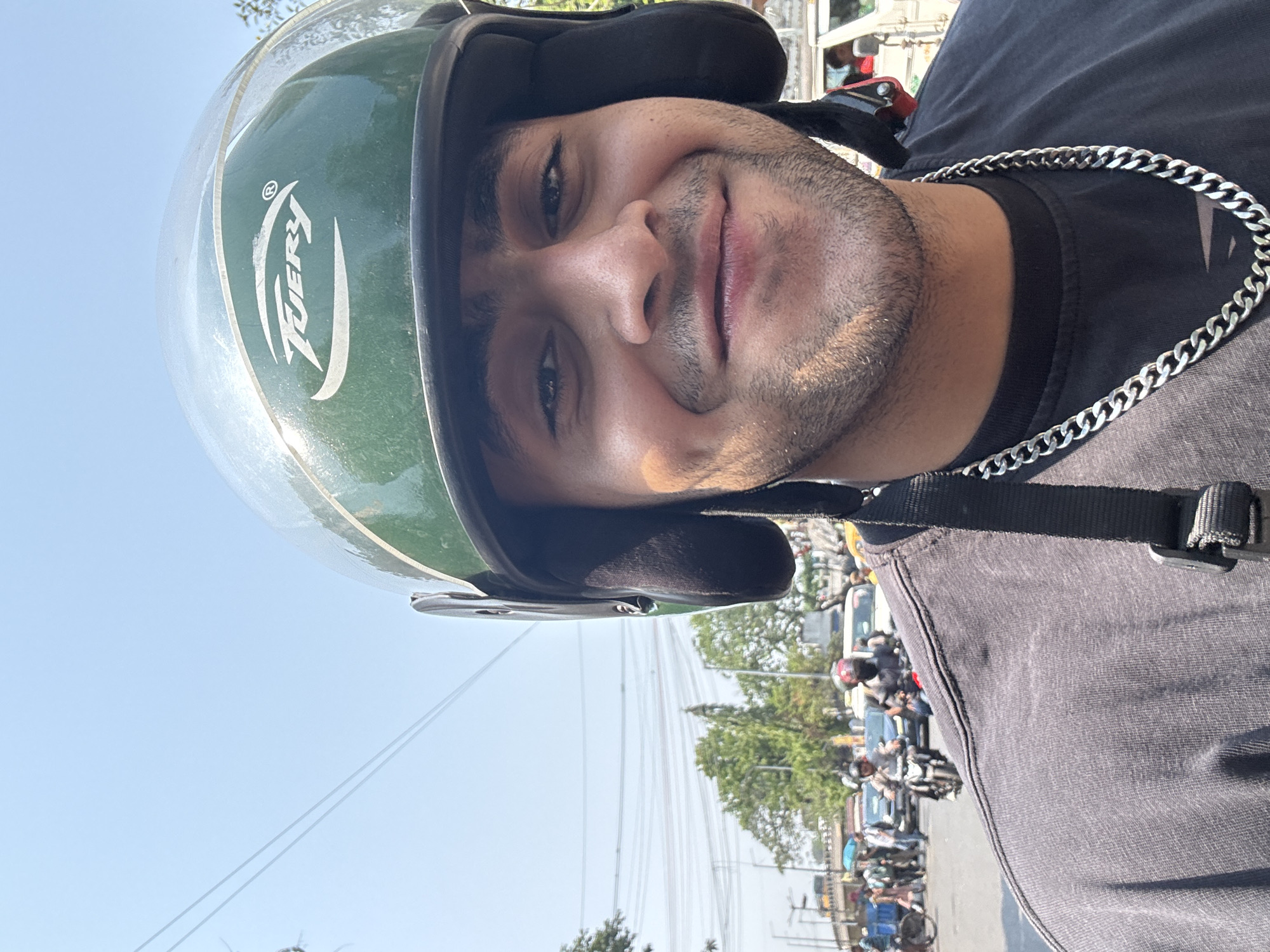 A man is seen wearing a green helmet with a clear visor, a black t-shirt, and a silver chain around his neck. He is smiling slightly at the camera. The background shows a busy street scene with cars, motorcycles, and trees, suggesting an urban environment. The presence of vehicles and infrastructure typical of South Asian cities indicates this location is likely within Kolkata, India, although specific identifying landmarks are not visible.