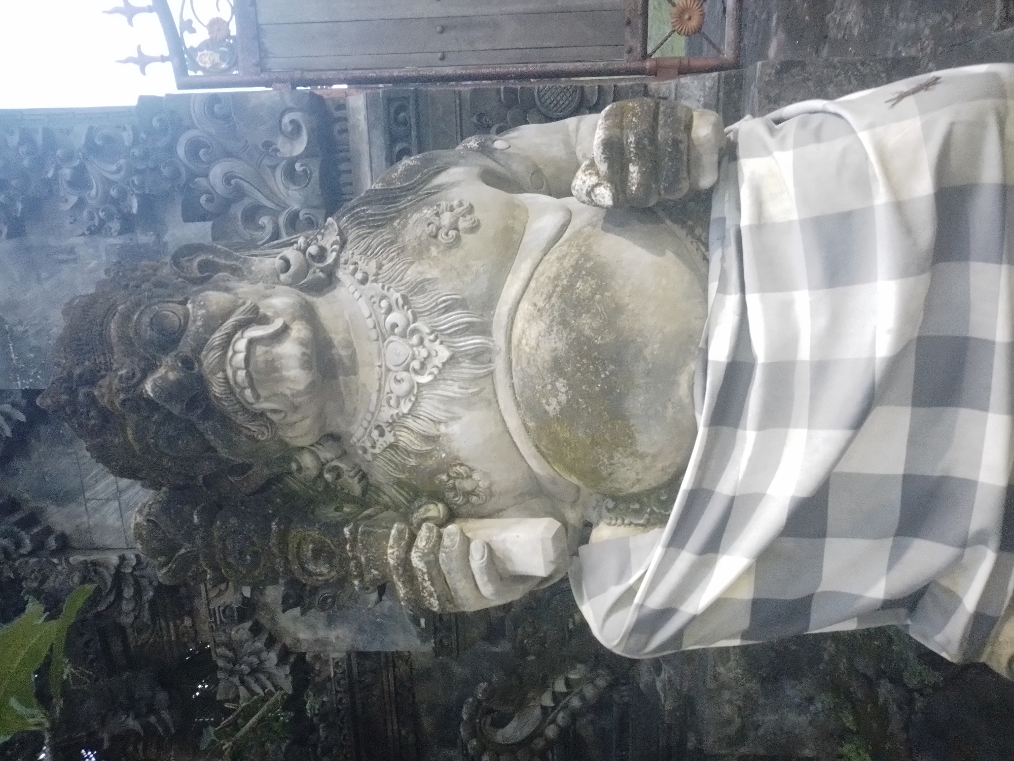 The image depicts a weathered stone statue of a demonic figure in Kuta Selatan, Indonesia. The statue is adorned with intricate carvings and traditional Balinese clothing in a grey and white check pattern is draped over the right side of its body. The figure has a menacing expression, with bulging eyes, a wide grin, and clenched fists. The surrounding environment appears to be a temple or a similarly religious setting, with ornate stone structures and foliage.