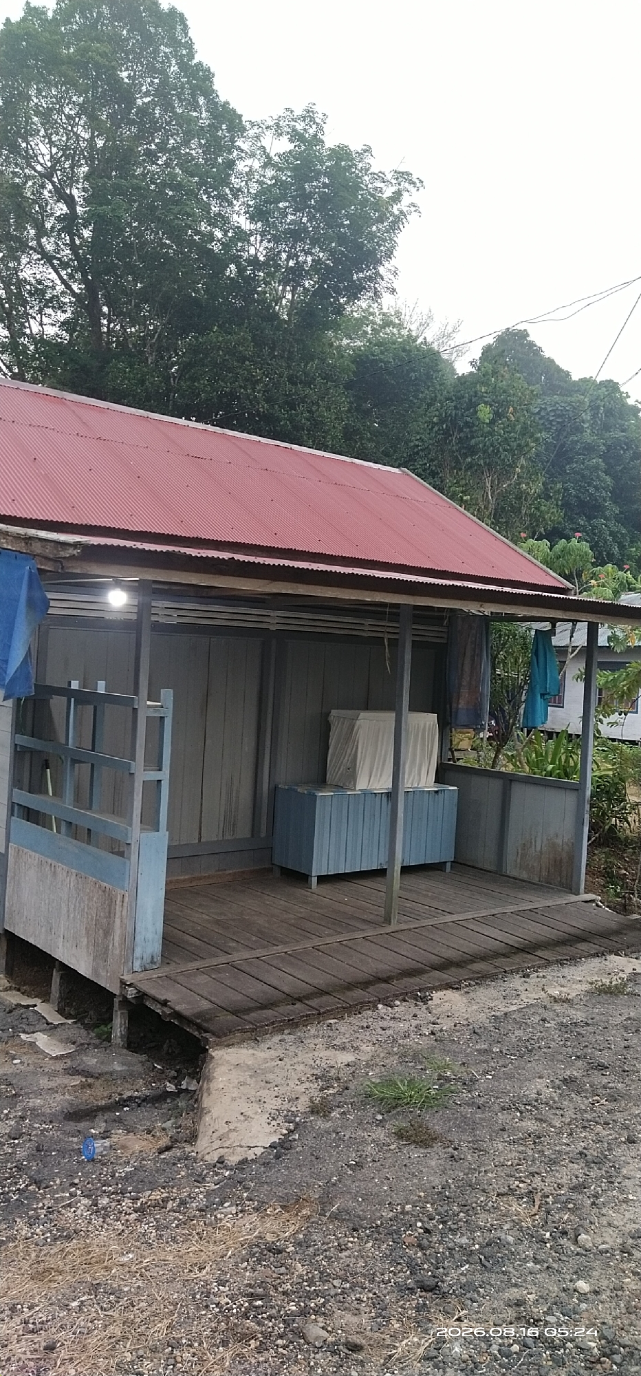 The image shows a small, weathered wooden structure with a red corrugated metal roof, located in the city of Muka Haji, Indonesia. The structure appears to be an open-sided porch or shelter, supported by simple wooden posts. The walls are made of vertical wooden planks painted a muted gray. A wooden deck extends from the front of the structure. Inside, a blue wooden cabinet is visible, covered with a white sheet or cloth. To the left, a blue tarp is draped over a railing. The ground in front of the structure is a mix of gravel and dirt, with some sparse dry grass. In the background, lush green trees and foliage are abundant, suggesting a tropical or sub-tropical environment. A white building with a dark roof is partially visible to the right, and power lines are present in the upper right corner. The sky is overcast, indicating a cloudy day. There are no people visible in the scene. The overall impression is one of a rustic, possibly abandoned or infrequently used, structure in a natural setting.