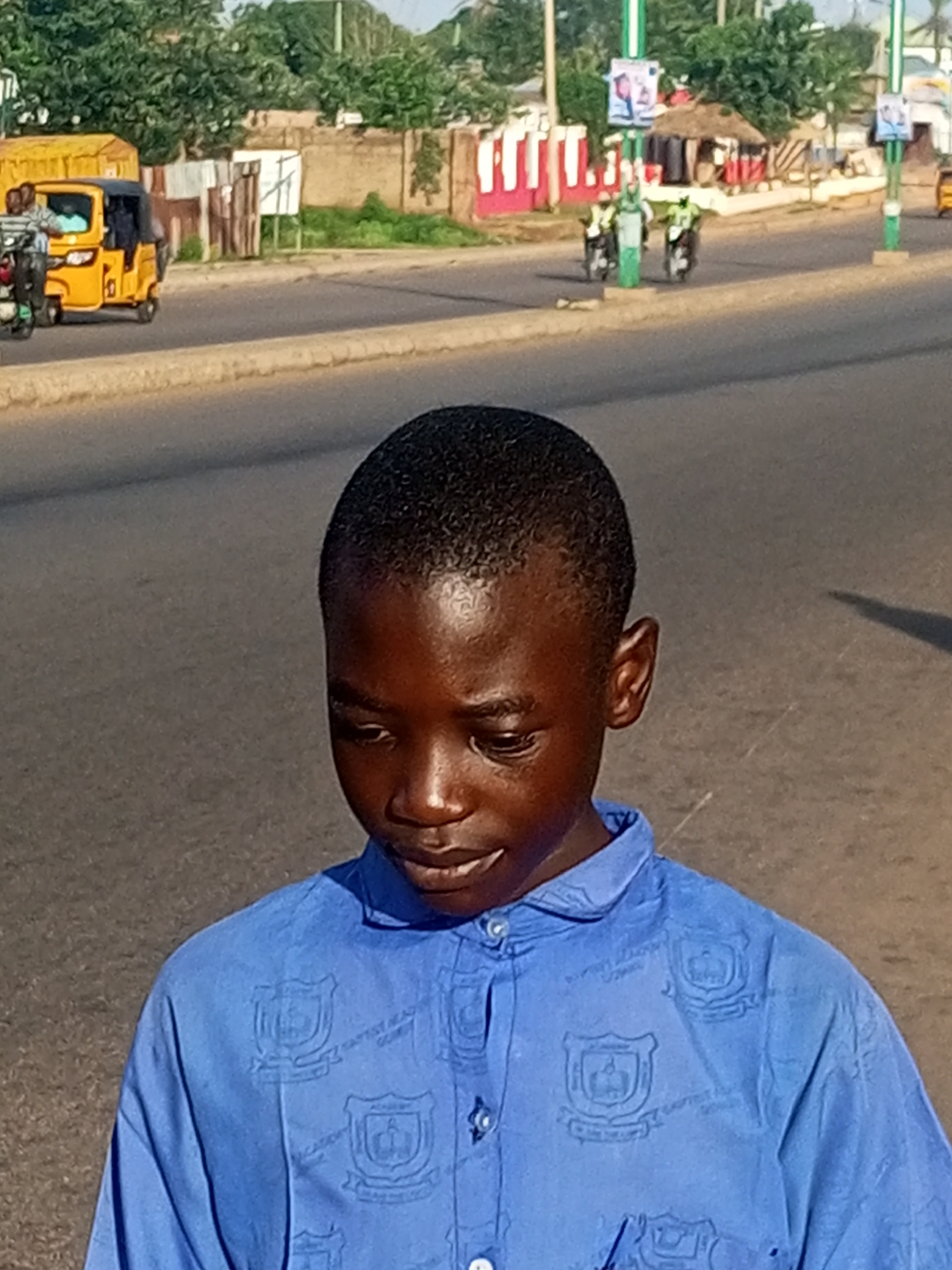 The image shows a young boy in a blue collared shirt standing on a street. The shirt has crests printed on it. The boy is looking down and to his right. In the background, there is a street with a yellow auto-rickshaw on the left and two motorcycles with riders in the middle of the frame. There are also trees, buildings, and a wall visible in the distance. The scene appears to be in an urban environment. Based on the context, this photo was likely taken in the city of Gombe, Nigeria, a place known for its auto-rickshaws and motorcycles being common modes of transportation.