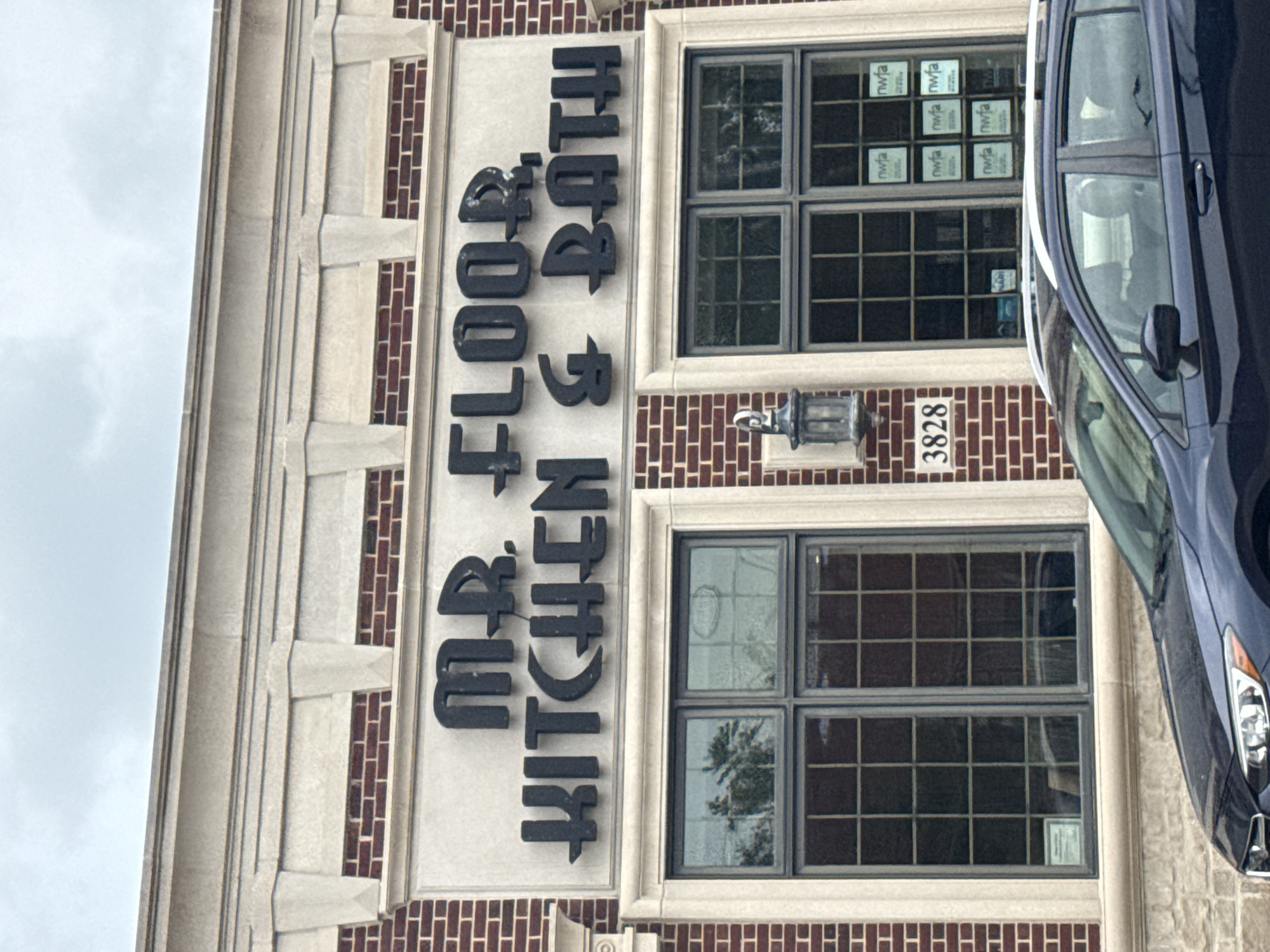 The image shows the exterior of a commercial building with the sign "MR. FLOOR KITCHEN & BATH" prominently displayed. The building is constructed with red brick and features decorative stone accents. There are large, multi-paned windows on the facade. A dark-colored car is parked in front of the building, partially obscuring the lower portion of the windows and the stone foundation. The scene appears to be on a street in Skokie, Illinois, United States of America. The sky is overcast. The building has a street number "3828" visible to the left of the windows.