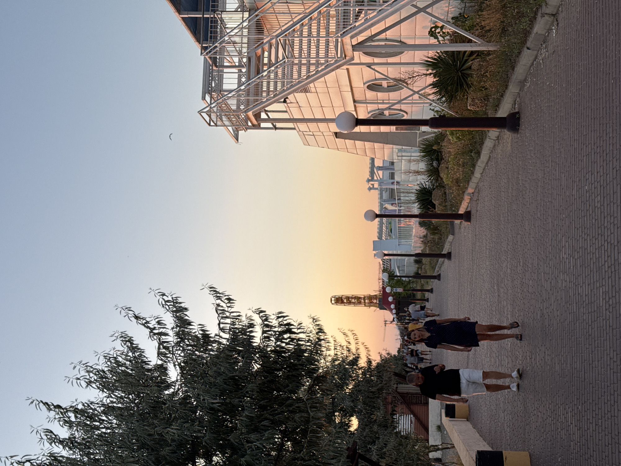 A daytime, eye-level shot captures a paved walkway in Novofedorovka, Ukraine, bordered by trees on the left and a modern building with outdoor stairs on the right. The sky transitions from a pale blue at the top to a warm orange hue near the horizon, indicating either sunrise or sunset. In the mid-ground, a Ferris wheel structure is visible, along with a row of streetlights lining the walkway. Several people are walking along the path, with a couple in the foreground. The man wears a black t-shirt and white shorts, while the woman is in a dark blue dress. The overall scene depicts a public space, likely a promenade or esplanade, during a pleasant time of day.