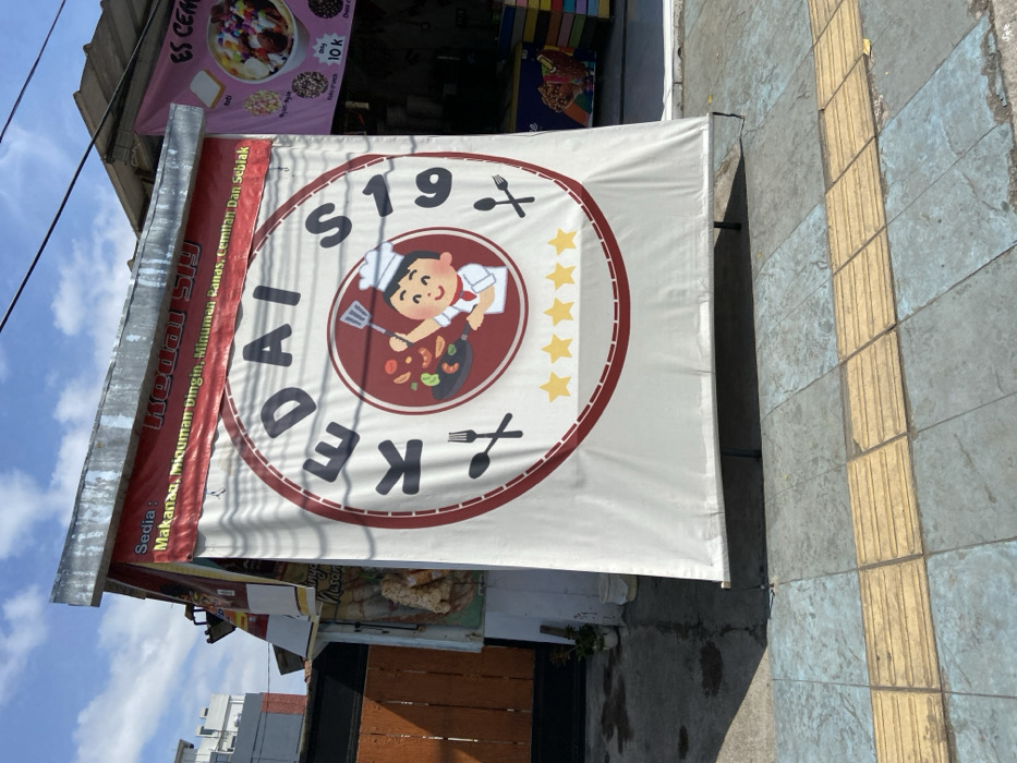 The image shows a street-side food stall in Padalarang, Indonesia, located at coordinates -6.85 latitude and 107.497 longitude. The stall is a small, white structure with a large white banner attached to its front. The banner features a red circle with the word "KEDAI" in large white letters and the number "S19" in white letters above and below the word. Inside the circle, there is an illustration of a smiling chef wearing a white toque and a red shirt, holding a spatula over a frying pan. The chef is standing on a red, circular surface. Five gold stars are centered below the circle. The banner is attached to the stall with black metal poles. In the background, there is a light-colored building with a dark-colored door and windows. To the right of the stall, there is another stall with a white sign featuring a colorful image of a bowl of ice cream. There is a concrete sidewalk in front of the stall with gray and yellow tiles. The stall has a sign that reads "Sedia: Makanan, Mmuaman Dingin, Minuman Panas, Cemilan Dan Seblak" in Indonesian. This translates to "We Serve: Food, Cold Beverages, Hot Beverages, Snacks and Seblak."