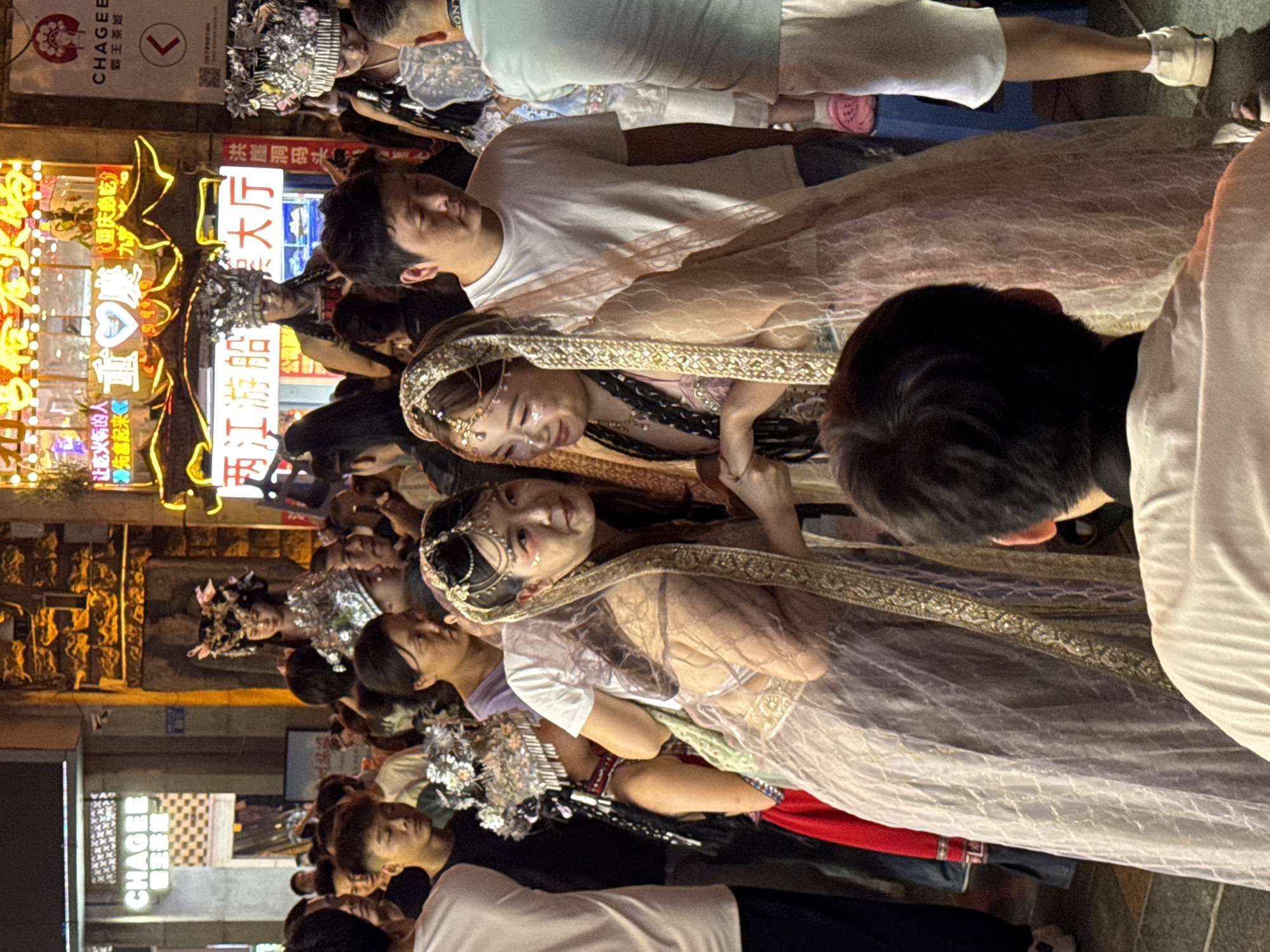 In Yuzhong District, China, a crowded night scene unfolds with individuals adorned in elaborate, traditional headwear and festive attire. Two women in the foreground, dressed in ornate, shimmering veils and headdresses, face each other and appear to be engaged in conversation or a performance. One woman has dramatic makeup applied around her eyes. Behind them, a diverse crowd of onlookers, dressed in casual, modern clothing, observes the scene. Various signs and neon lights, some in Chinese characters, illuminate the background, suggesting a vibrant street or marketplace. The overall impression is of a cultural event or celebration taking place in a bustling urban environment.