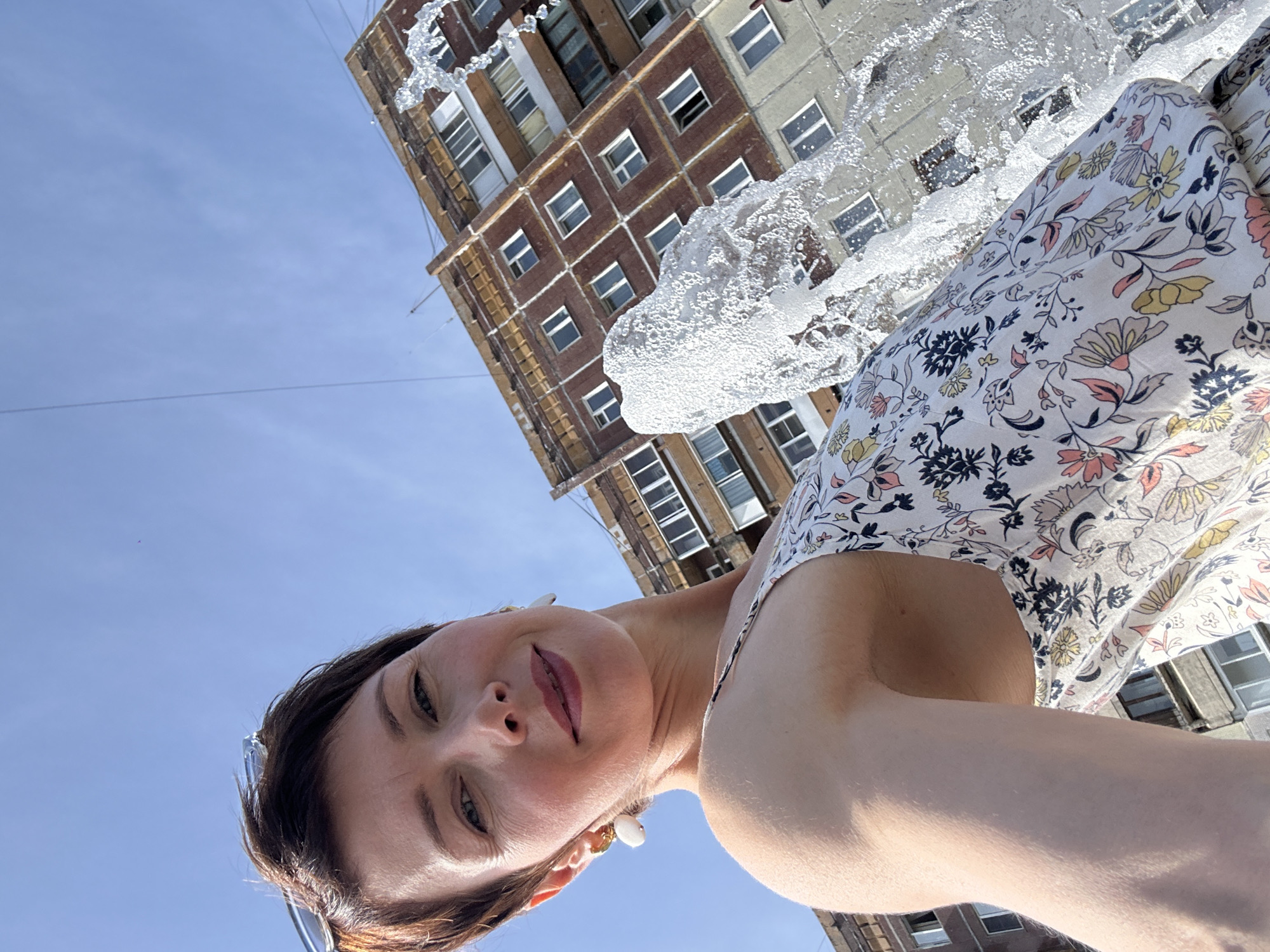 A woman with short brown hair and large white earrings takes a selfie outdoors in front of a multi-story building. She is wearing a light-colored dress with a floral pattern. A fountain sprays water to her right. The building, with its brick and concrete facade, appears to be an apartment complex located in Kemerovo, Russia. The sky is clear and blue.
