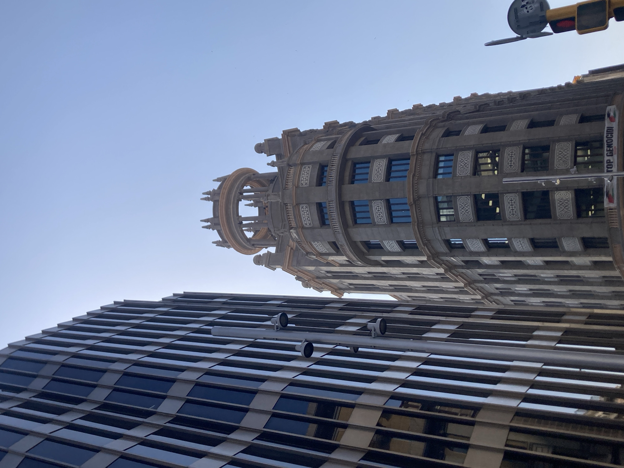 In Barcelona, Spain, the image captures a low-angle view of two distinct architectural styles. On the left, a modern skyscraper with a facade of dark, vertical glass panels dominates the foreground. To its right, a more ornate, older building stands tall, featuring rounded corners, decorative stone carvings, and arched windows. The sky above is clear and bright blue. A streetlamp with multiple light fixtures is visible attached to the side of the modern building. At the base of the older building, a banner with text that appears to read "TOP GENOCIDI" is visible, along with traffic lights and street signage to the far right. No people are clearly discernible in the frame.