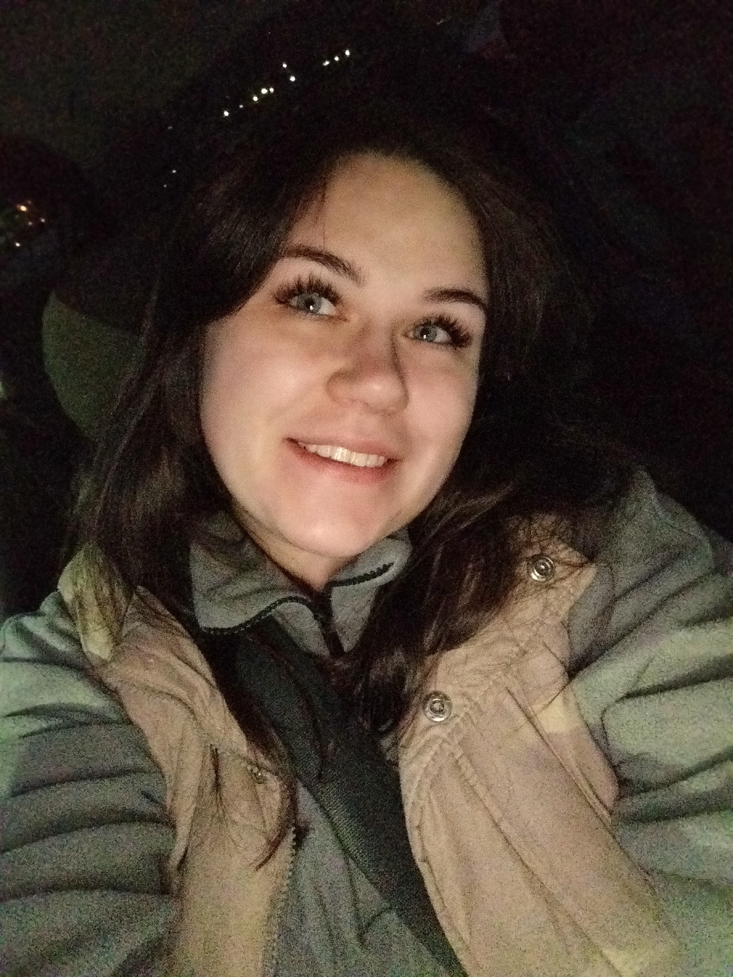 The image displays a young adult female in the city of Chaikovsky, Russia, seated in what appears to be a vehicle at night. She has fair skin, dark brown, wavy hair, and light-colored eyes with long, dark eyelashes. Her expression is a wide smile, revealing white teeth, and her gaze is directed slightly upwards and to her right. The subject wears a grey zippered garment, underneath a beige quilted vest with visible metallic snap fasteners. A dark strap, consistent with a seatbelt, crosses her chest diagonally. The background is dark, with indistinct shapes suggesting a vehicle interior, and scattered small, blurry white lights are visible in the upper left, indicative of distant urban lighting at night. The primary illumination source lights the subject's face from the front.