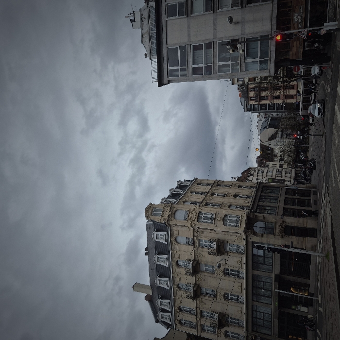 The image depicts a multi-directional urban intersection at Place de l'Homme de Fer, Strasbourg, France, under a heavily overcast grey sky.
On the left, a prominent multi-story historic building features a light-colored, ornate facade with decorative balconies and a dark grey Mansard roof punctuated by multiple dormer windows. The ground floor displays large glass windows and signage, including a green "MONOPRIX" logo.
To the right, a multi-story modernist building with a light grey concrete facade and large, dark-framed rectangular windows stands opposite the ornate structure. Its flat roof shows visible equipment. A red traffic light is prominently displayed at its corner, directing traffic at the intersection.
The foreground consists of an asphalt road marked with white pedestrian crosswalks. Several vehicles are present: a silver car is positioned near the center of the intersection, and other vehicles are partially visible further down the streets. Bicycles are parked near the base of the ornate building. Overhead power lines traverse the sky between the buildings. Multiple pedestrians are visible on sidewalks and near crosswalks, with one figure wearing a yellow jacket near the left building. Streetlights and poles are integrated into the urban infrastructure. The overall environment is one of an active city street during daytime, characterized by dull lighting due to the thick cloud cover.