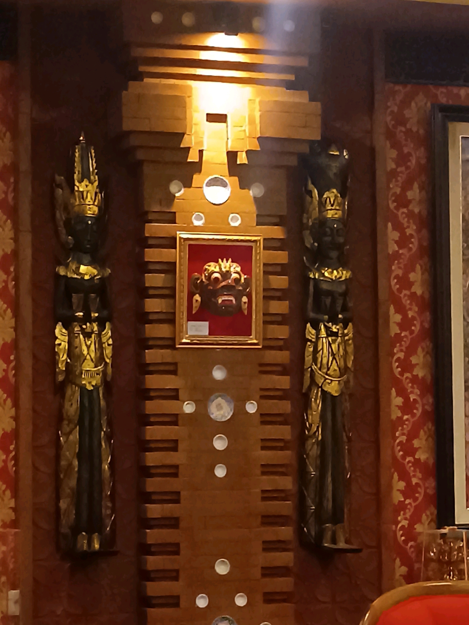 The image depicts an ornate interior decoration featuring two tall, dark statues with gold accents, flanking a central wooden structure. The central structure has a tiered design and houses a framed mask with a fierce expression. The walls are covered in a red and gold damask-patterned wallpaper. The lighting is warm, with some bright spots suggesting overhead lights. The scene appears to be in a dimly lit, indoor environment, possibly a reception area or a ceremonial space. The style of the statues and mask suggests an Indonesian or Southeast Asian cultural influence, potentially Balinese, given the reference to Tabanan, Indonesia.