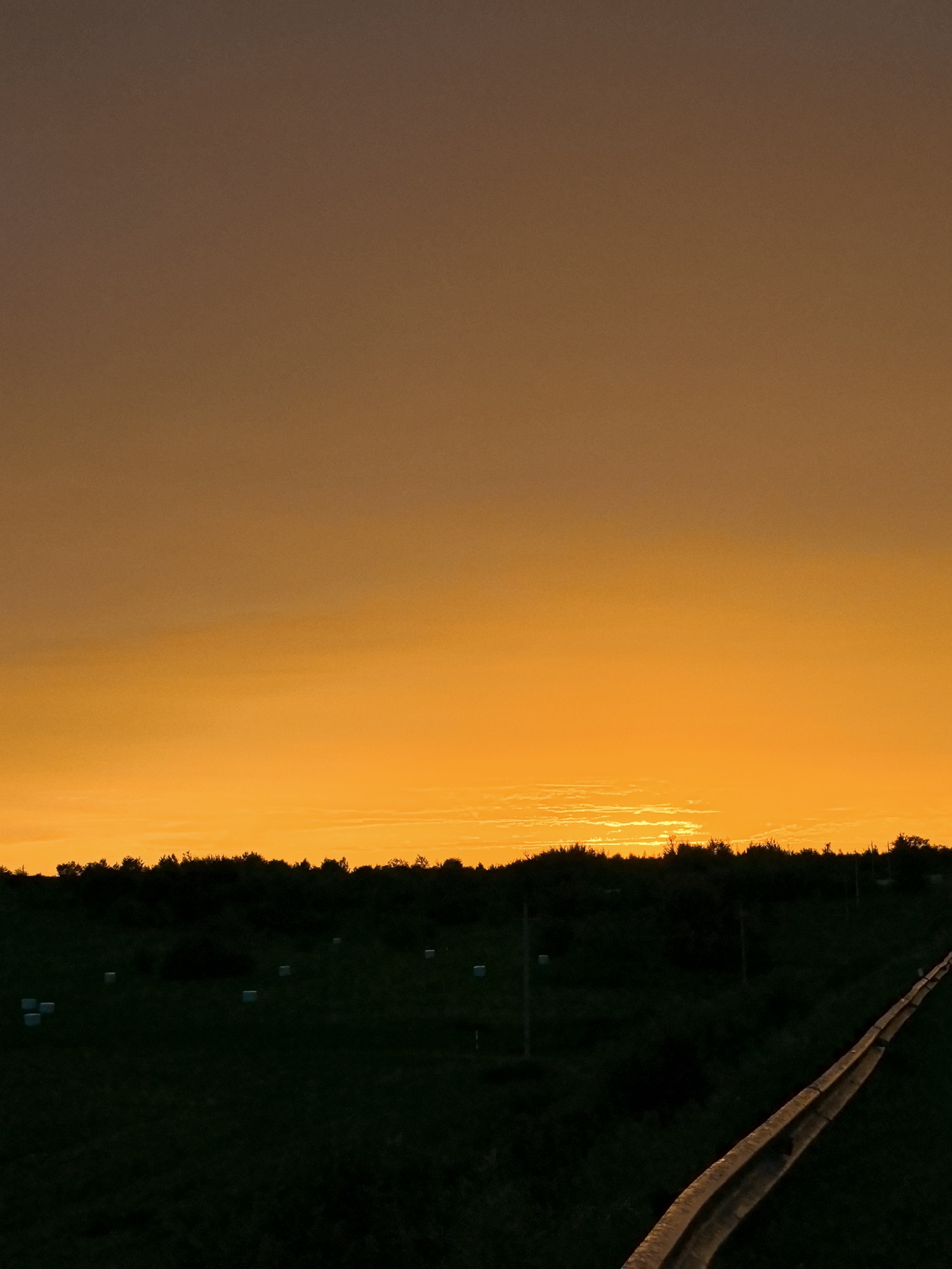 A dark silhouette of trees forms the horizon line of a rural landscape under a vibrant orange sunset. The sky transitions from a deep brown at the top to a bright, fiery orange near the horizon, with wispy clouds visible. In the foreground, a dark, grassy field is dotted with several white bales of hay and a few utility poles. A guardrail runs diagonally across the right side of the image, also silhouetted against the dim light. The scene is quiet and still, evoking a sense of peace at the end of the day.