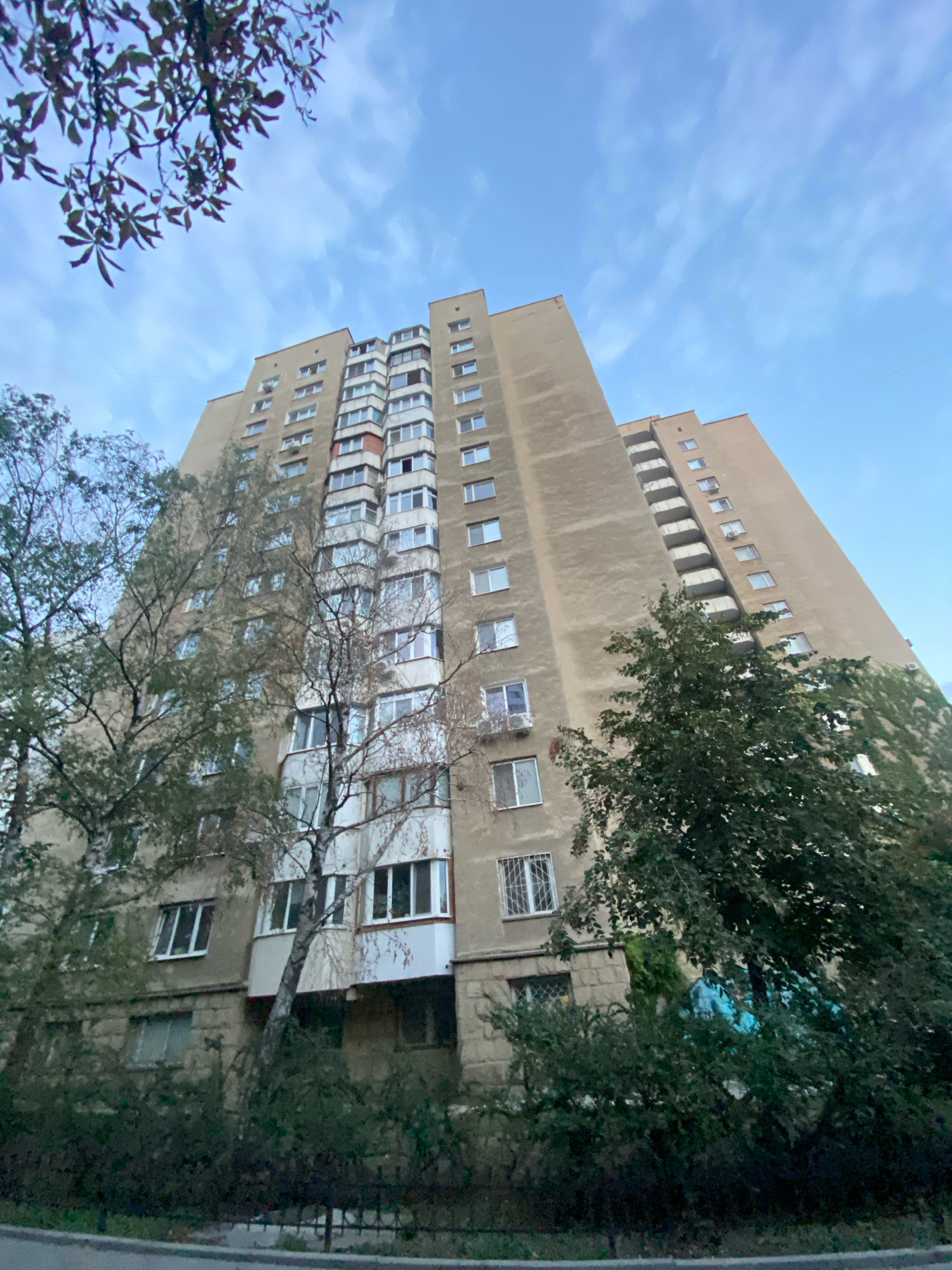 A low-angle, wide shot captures a tall, concrete apartment building against a cloudy, pale blue sky. The building is light brown, with numerous windows and balconies. Trees with green and white foliage flank the building, with some branches extending into the frame from the top left. A dark green hedge and a black metal fence are visible at the base of the building. The scene appears to be a residential area.