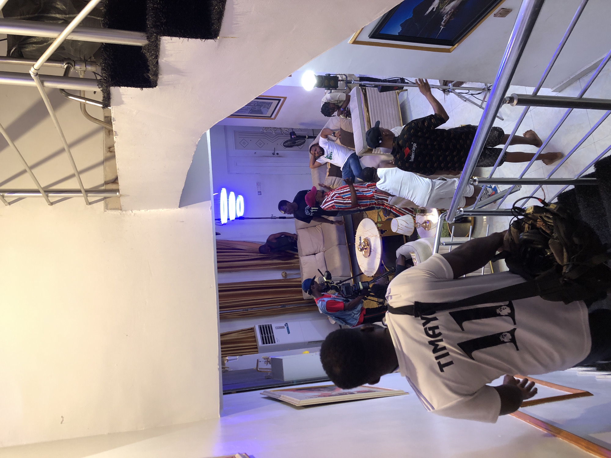 The image captures an indoor scene from an elevated perspective, looking down into what appears to be a living room or studio space where a production, likely a photoshoot or video shoot, is underway.
In the immediate foreground, the back of a person wearing a white t-shirt with "TIMGAY" and the number "11" printed in black is visible. This person also has a camera or equipment bag strapped to their back and is standing near a white staircase with black, textured steps and metallic railings.
Below, in the main room, several individuals are present. One person in a black patterned shirt with "Balenciaga" text is standing barefoot near a metallic railing, looking towards the activity. Another person is seen adjusting a bright studio light on a stand. Further back, individuals are seated or standing around sofas and tables. One person is operating a camera on a tripod, focusing on the scene.
The setting features light-colored tiled floors, white walls, and elegant brown curtains. Decorative elements include framed artwork, a modern floor lamp, and a series of prominent blue neon lights. An air conditioning unit is visible on a far wall. The furniture consists of light-colored sofas and round tables, one with a reflective, possibly metallic, surface. The scene is well-lit, primarily by artificial light sources, indicating an event taking place regardless of the time of day. No strong emotions are discernible, with individuals appearing focused on the activity.