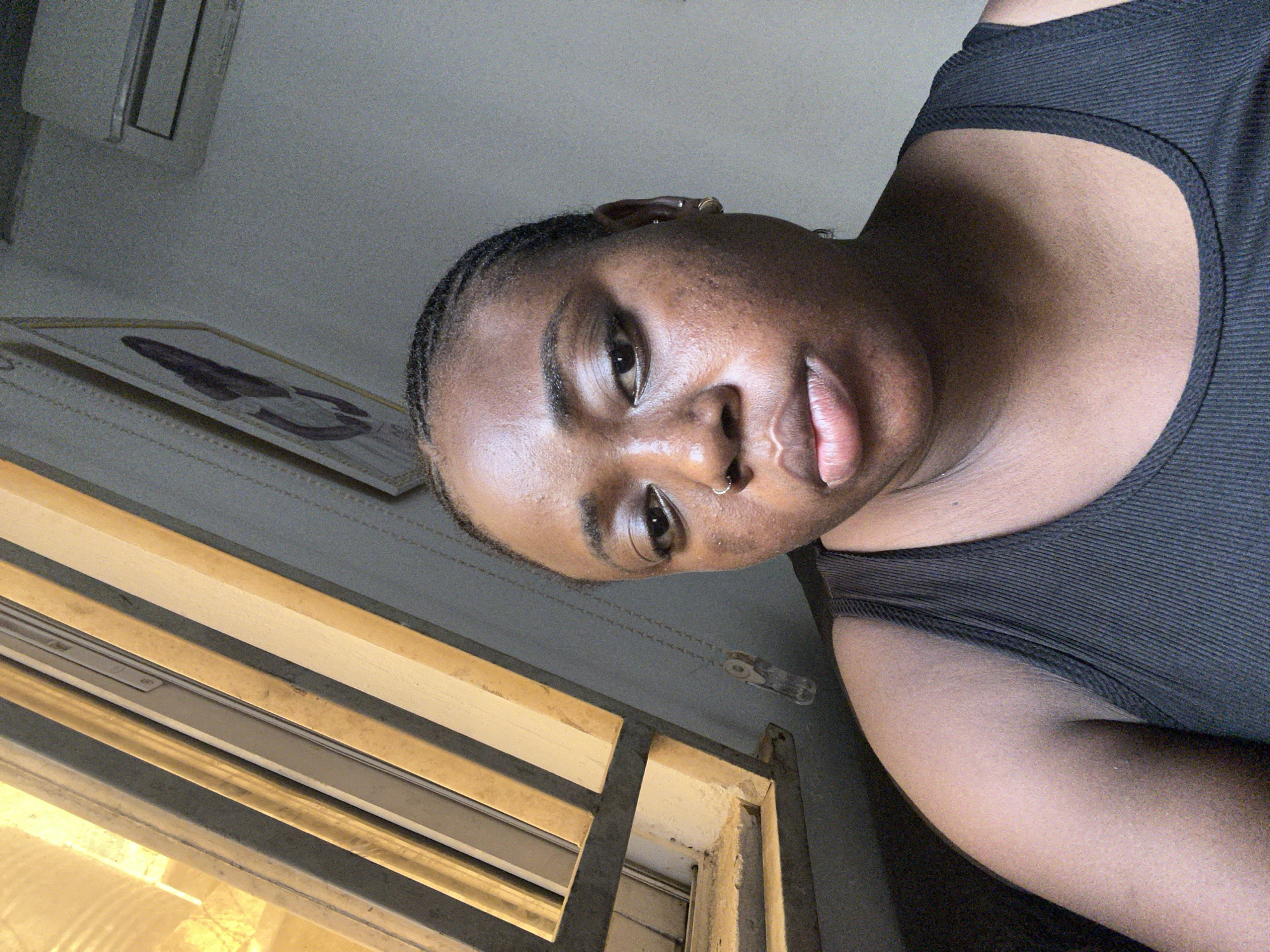 A close-up, slightly tilted, indoor portrait of a woman with dark skin wearing a black tank top. She has short, braided hair and a nose ring. Her gaze is directed slightly downwards. Behind her is a window with horizontal blinds. In the upper left corner is an air conditioning unit, and below it, a framed picture hangs on the wall. Based on the visual details available, the location cannot be pinpointed to Abuja, Nigeria. Further contextual details or specific landmarks would be required for precise identification.