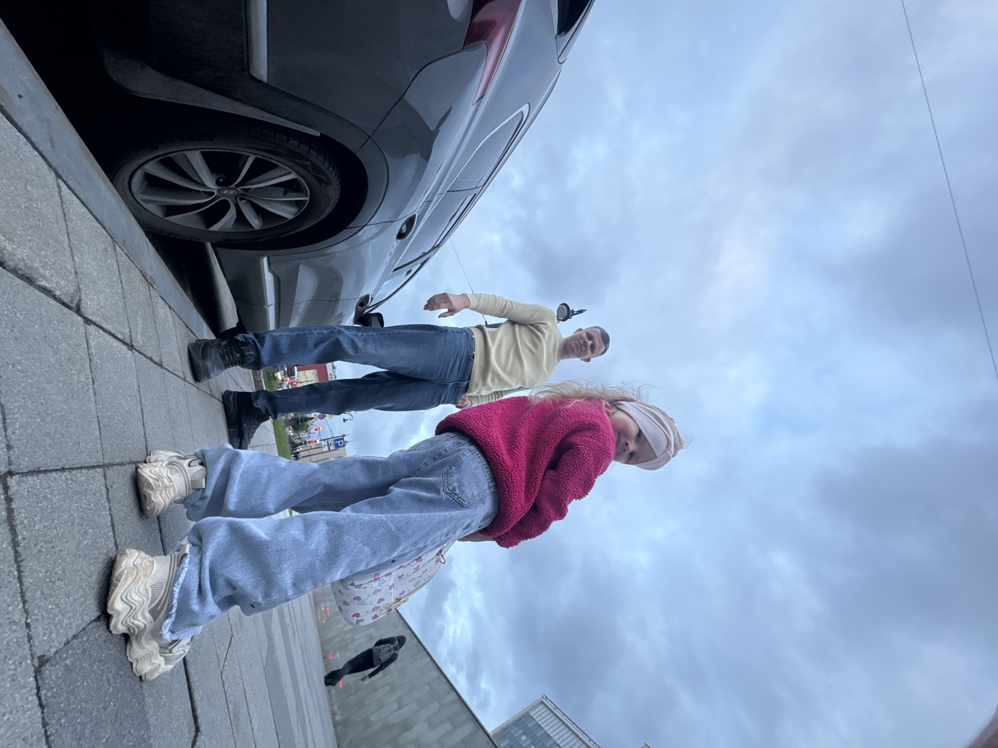 A low-angle perspective captures a man and a young girl standing beside a gray car on a city street. The girl, in the foreground, wears a red fleece jacket, light blue jeans, and chunky sneakers. She has a white headband and carries a white bag. The man behind her is dressed in a light yellow sweater and blue jeans. The car is parked close to the curb. The background features a cloudy sky, a glimpse of cityscape, and another pedestrian walking on the sidewalk. The location is Perm, Russia.