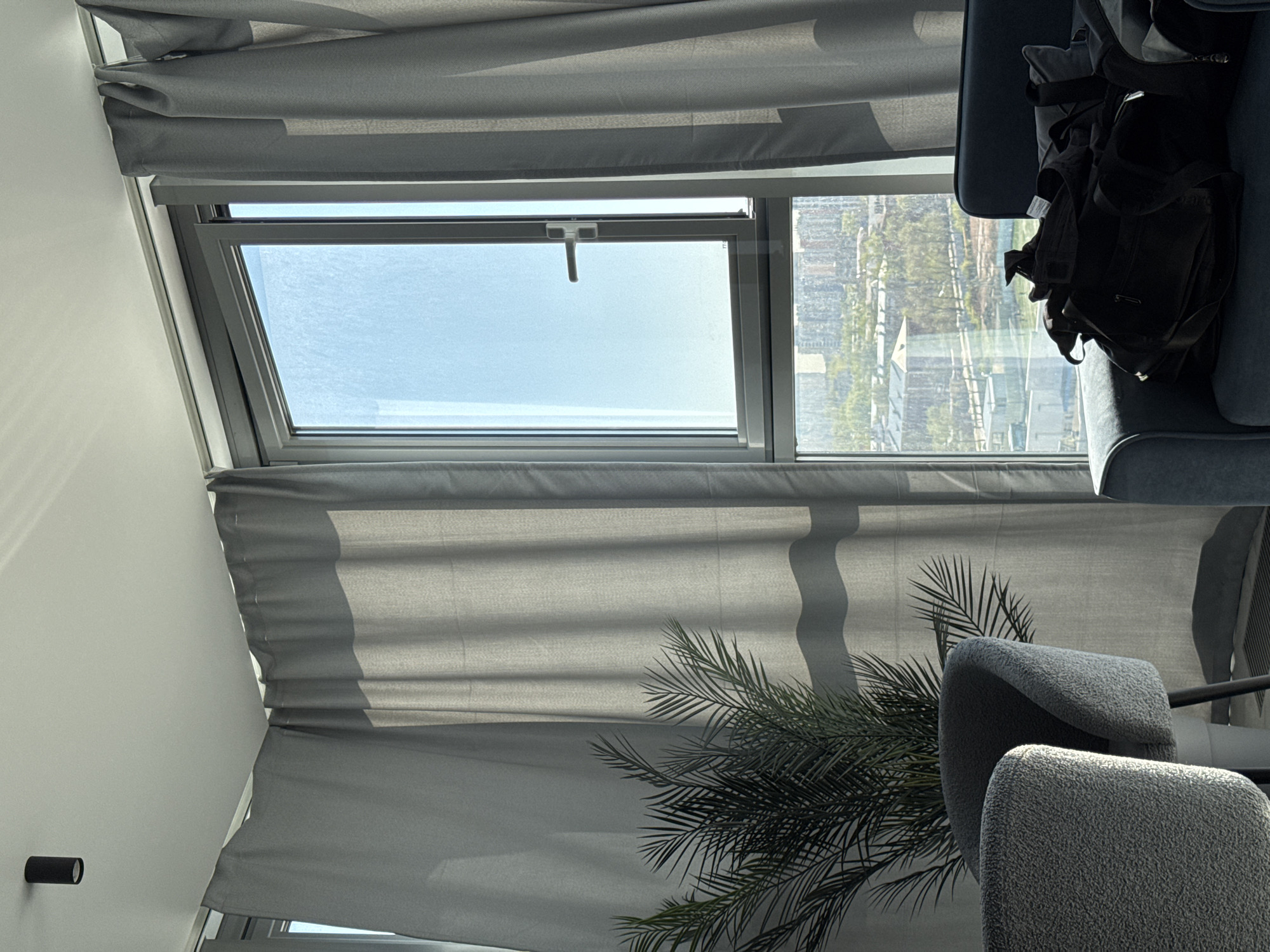 The image shows an interior setting featuring a window with gray curtains, partly obscuring the view outside. The window is a top-hinged type and is slightly open, revealing a clear sky. In the background, through the window, a cityscape can be seen, consisting of tall buildings and greenery. In the foreground, parts of gray chairs are visible, along with a potted plant placed near the window. A black bag rests on a blue armchair, and a small black cylindrical object is fixed on the wall. The scene is bathed in natural light.