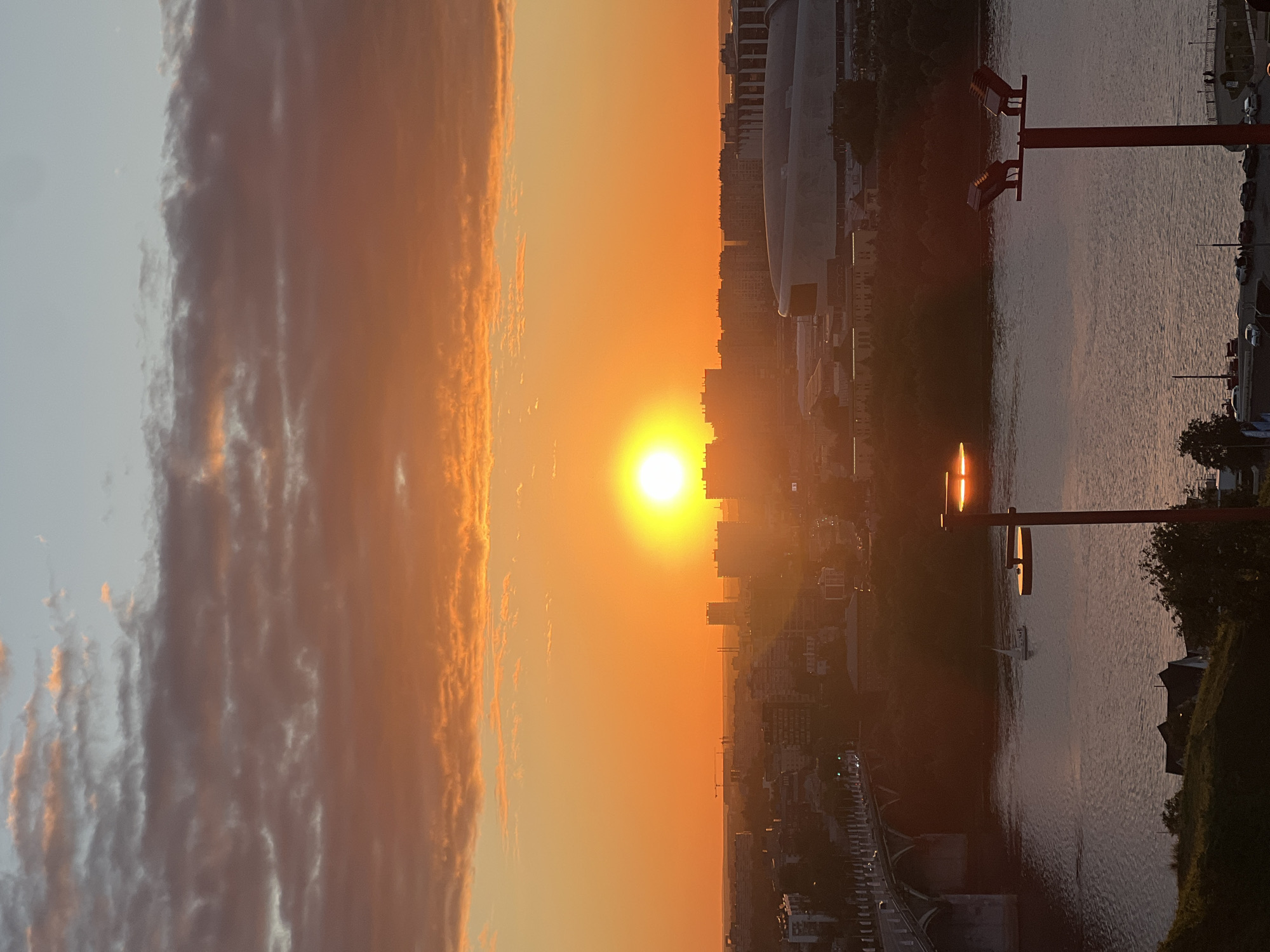 This is a wide-angle, vertical, full shot taken during sunset from a high vantage point overlooking the city of Nizhny Novgorod, Russia. The sun, a bright orange orb, is the dominant feature in the sky, casting a warm glow over the city and clouds. The sky is a gradient of orange and blue, with scattered clouds tinged orange and pink.
In the middle ground, a river, possibly the Volga, flows through the city. A white sailboat is visible on the water. A bridge with multiple arches spans the river on the left side of the frame. The city skyline features numerous buildings of varying heights and architectural styles, including a modern stadium-like structure with a curved roof in the mid-right. Silhouettes of trees line the riverbanks.
In the foreground, on the right, a modern streetlamp with two square lights on a red pole stands prominently. On the left, a grassy slope is visible, with some houses and trees at its base. The overall scene depicts a serene urban landscape bathed in the warm light of a sunset. There are no visible people in the image.