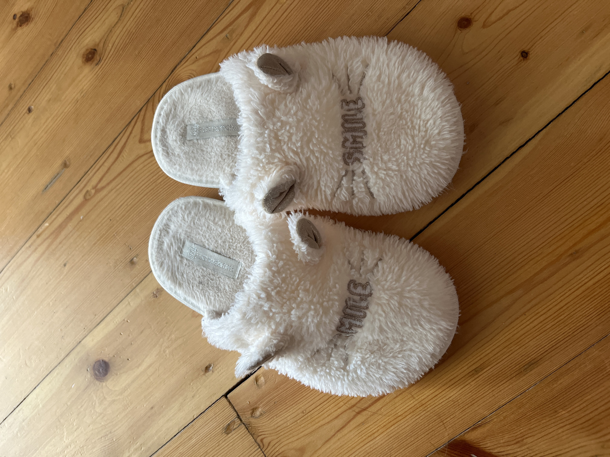 A pair of fluffy white slippers with grey embroidered "smile" text and small grey bunny ears sit on a light brown wooden floor. The slippers are positioned next to each other, slightly angled. The floorboards are wide and show visible grain and knots. The slippers are the sole objects in the image, and the lighting suggests an indoor environment.