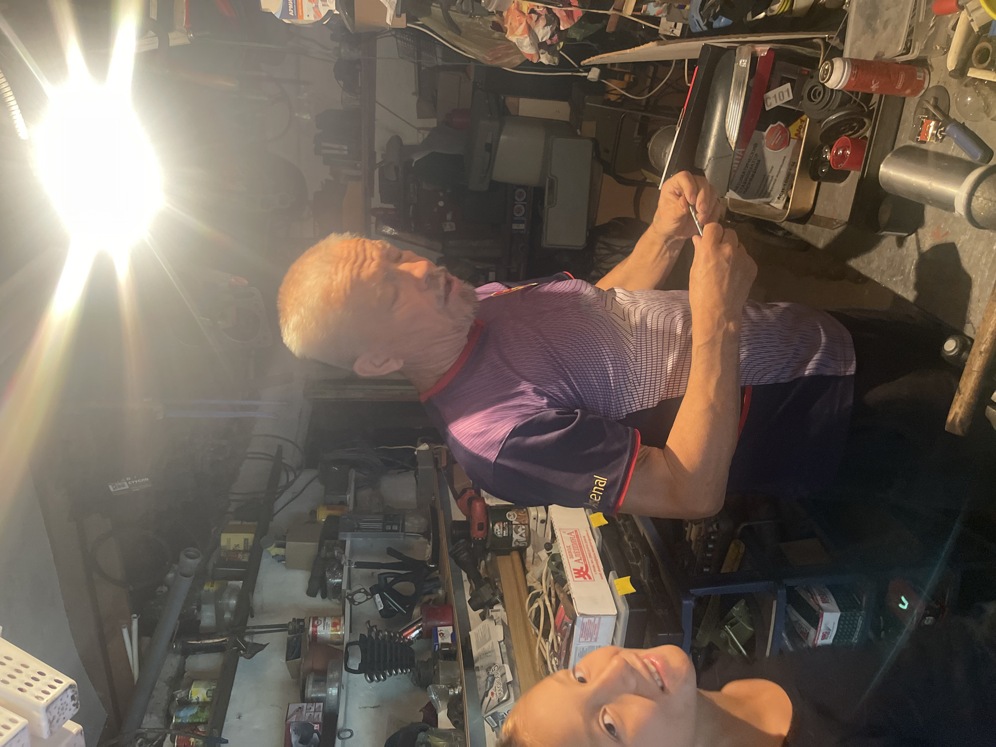 In a cluttered garage workshop located in Sibirskiy, Russia, an older man with grey hair and a goatee, wearing a purple and black patterned t-shirt, is actively engaged in a task involving a small metallic object, possibly a screw or a nail, which he holds in his right hand. He appears to be inspecting or manipulating this item. To his left, a young girl with blonde hair and a bright smile is partially visible, looking towards the camera. She is wearing a dark t-shirt. The garage is filled with shelves laden with various tools, cans, and other items, indicative of a working space. A bright overhead light illuminates the scene.