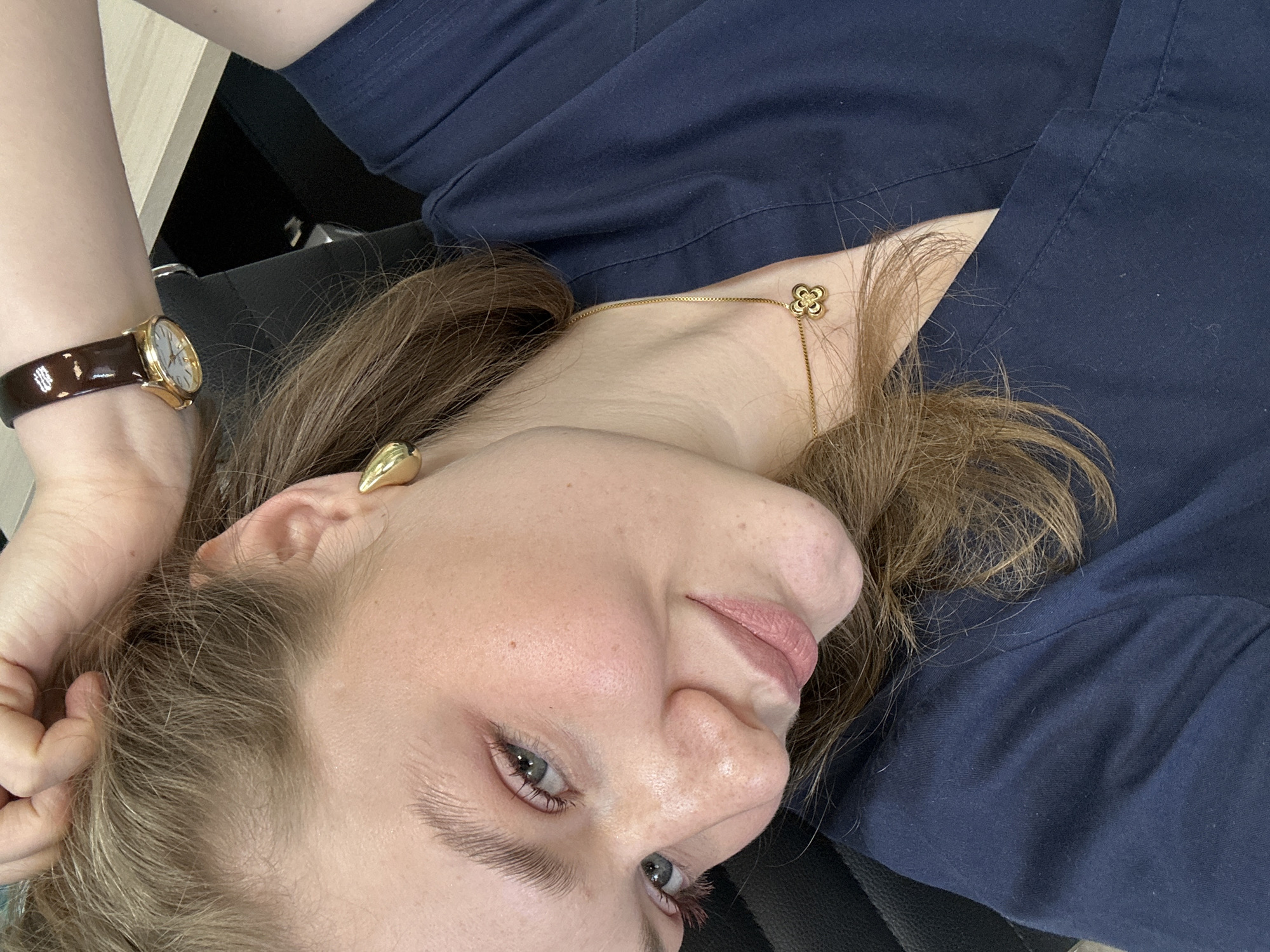A close-up, tilted view of a woman with fair skin and light brown hair. She is wearing a dark blue top. Her head is resting on her hand, which has a brown leather watch with a gold face. She has gold teardrop-shaped earrings and a gold necklace with a floral pendant. She has light blue eyes. The background includes a black chair and a light-colored table. There is no information in the image to suggest it was taken in Kazan, Russia.