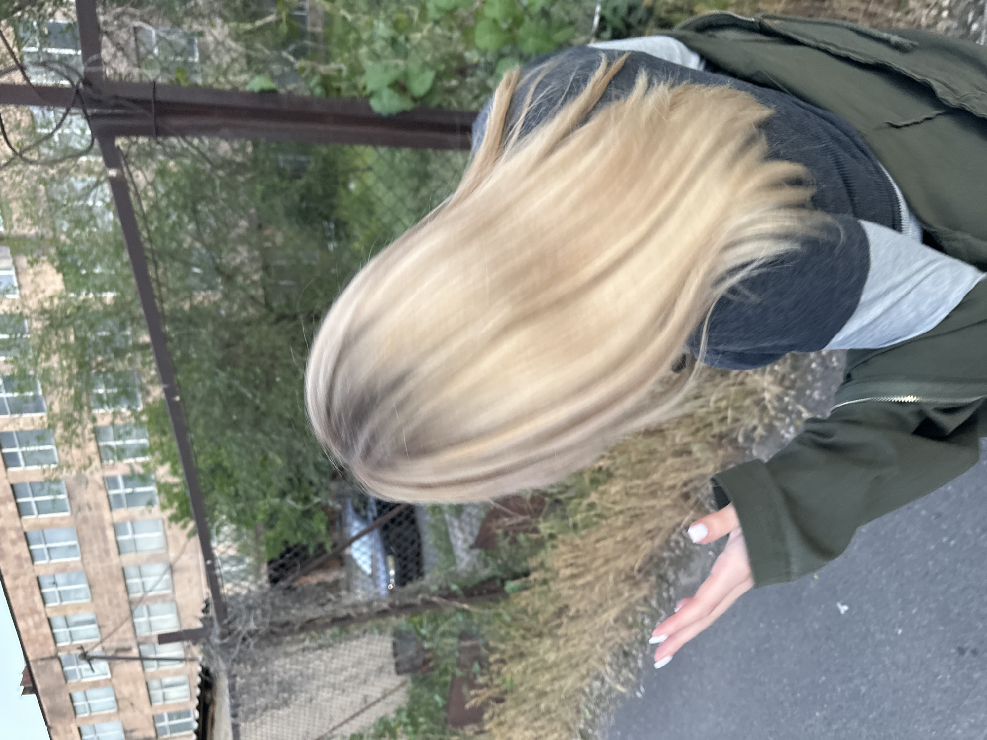A tilted, medium shot depicts a blonde-haired person outdoors in Yerevan, Armenia. The person is wearing a dark grey top, a light grey shirt beneath it, and an olive green jacket. Their left hand, with white painted nails, is extended towards the bottom right corner of the frame. A dilapidated metal fence runs vertically along the left side, separating the person from a multi-story building with numerous windows in the background. Sparse vegetation is visible between the fence and the person, with asphalt ground occupying the lower right portion of the image. The background reveals hints of trees and foliage.