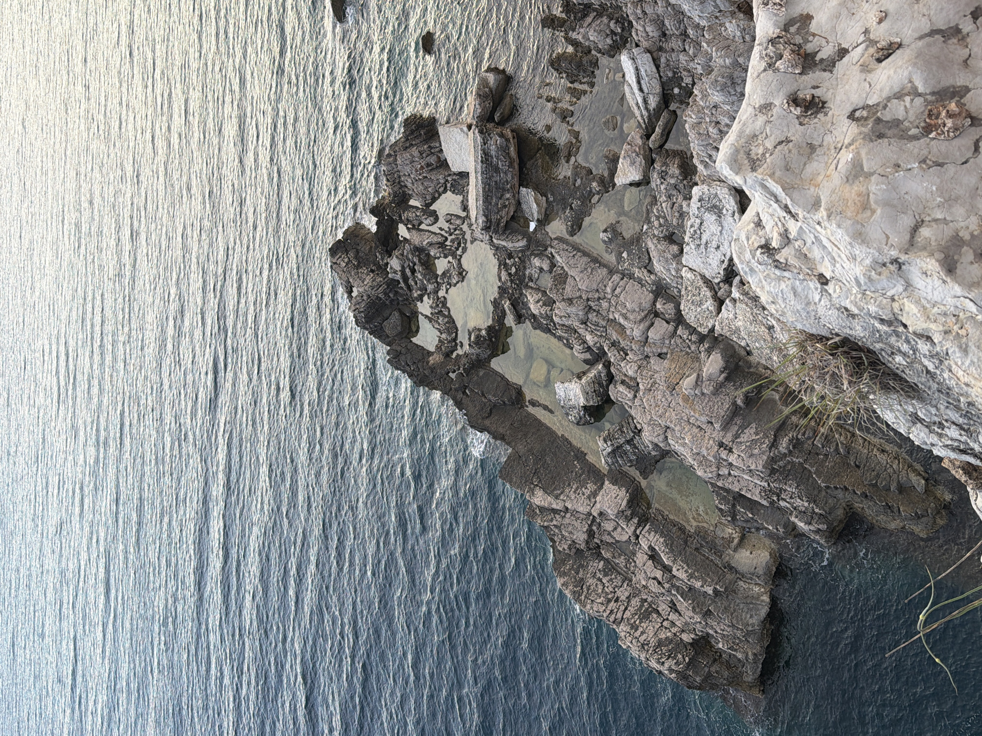 The image shows a rocky coastline with the Adriatic Sea in Mirište, Montenegro. The foreground features large, weathered rocks with visible textures and variations in color, ranging from light grey to darker shades of brown and black. Some rocks are partially submerged in the clear, shallow water that has collected in natural pools among the formations. The water itself is a deep blue-green, with small ripples reflecting the ambient light. Beyond the immediate rocky area, the sea extends to the horizon, with a lighter, more reflective surface suggesting a gentle breeze. The scene is natural and undisturbed, with no people or man-made objects visible, except for possibly some very distant, indistinct structures on the horizon, not clearly identifiable.