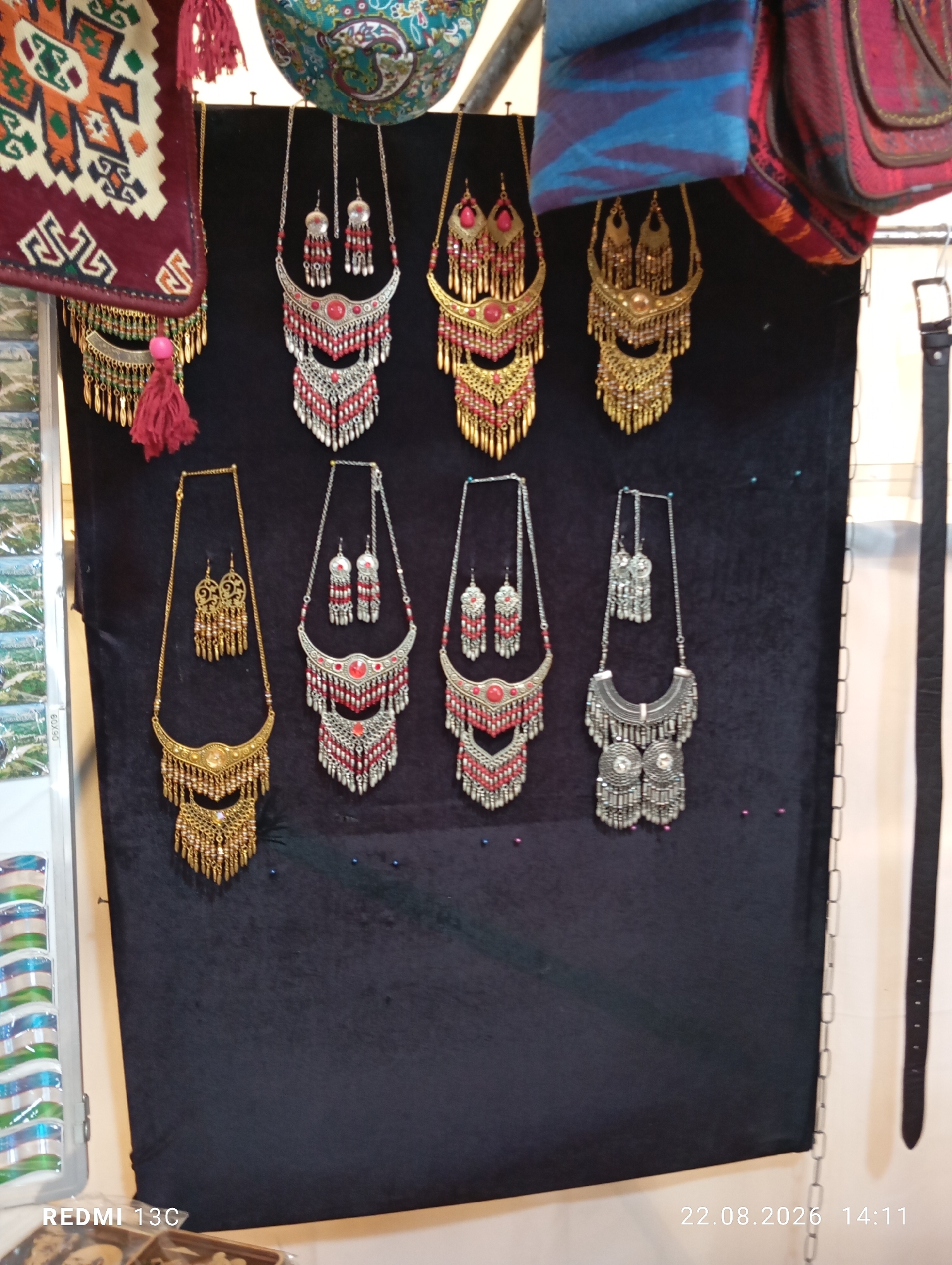 The image shows a display of traditional Uzbek jewelry, likely in a market stall or shop. The jewelry is predominantly necklaces and matching earrings, featuring intricate metalwork, beads, and colorful accents. The pieces are hung against a dark fabric backdrop. In the upper part of the image, there are also textile items, possibly bags or decorative cloths, with vibrant patterns. A black belt hangs to the right. The bottom left corner displays a sticker with "REDMI 13C" and the bottom right shows a date and time: "22.08.2026 14:11". While the specific location in Amirsay, Uzbekistan is not explicitly visible, the style of jewelry and textiles strongly suggests this region, known for its traditional crafts.