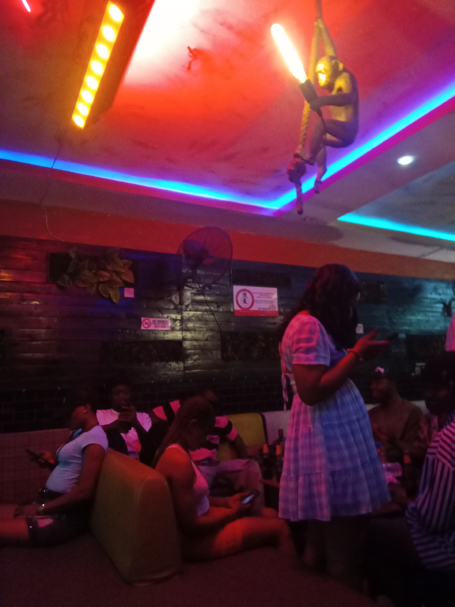 The scene is an indoor setting, likely a bar or lounge, in Obosi, Nigeria. Several people are present, some seated on a couch. One woman is standing, wearing a light-colored plaid dress and looking at a mobile phone. Other individuals are seated or reclining on the couch. The lighting is colorful, with neon lights on the ceiling. A decorative sculpture of a monkey hangs from the ceiling, holding a light. A fan is mounted on the wall, along with a "no smoking" sign. The overall atmosphere appears dimly lit and relaxed.